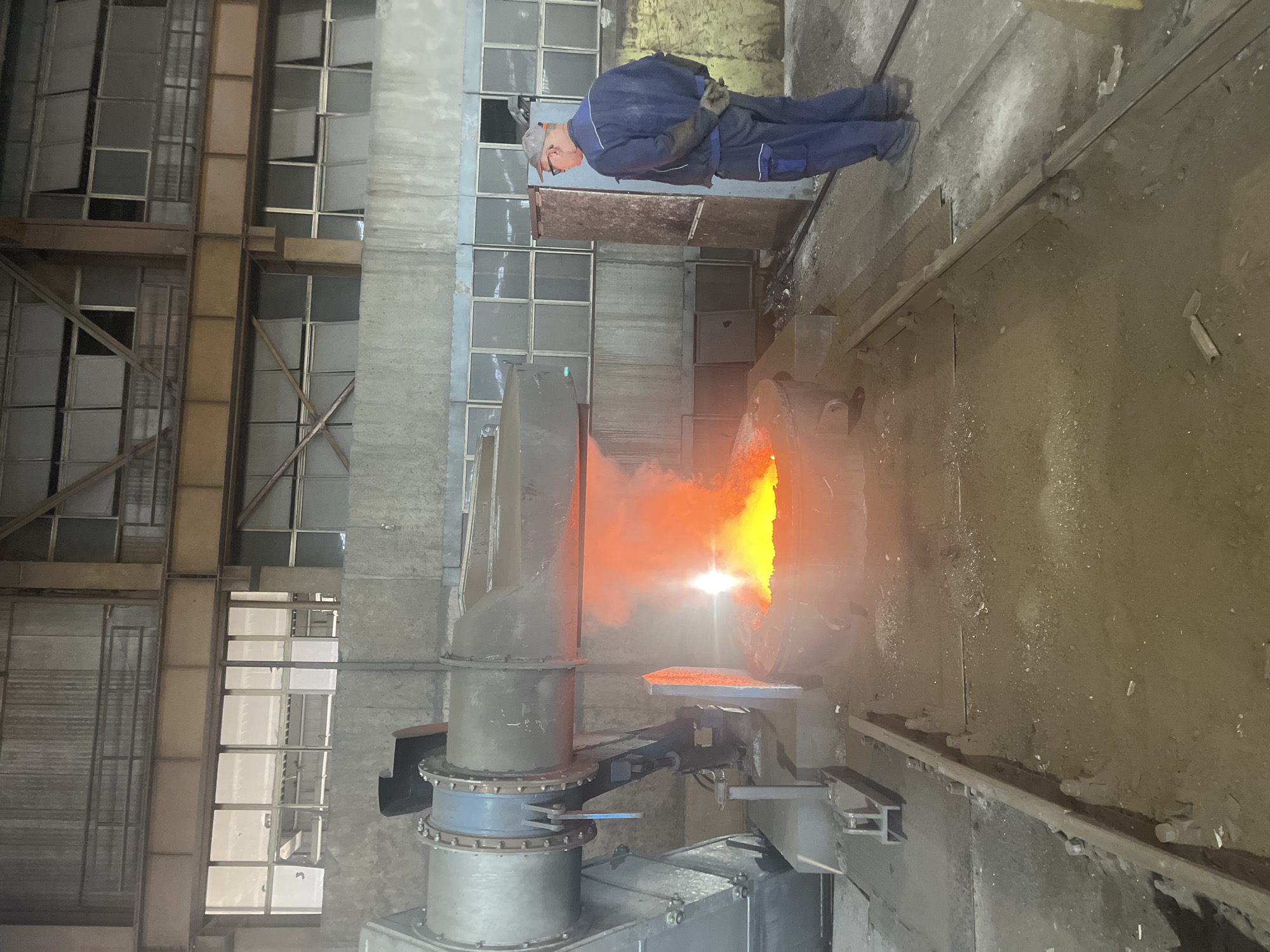 The image depicts an industrial setting, possibly a foundry or metalworking factory, located in Plovdiv, Bulgaria. A worker wearing a blue jumpsuit, gloves, and a cap stands near a large piece of industrial machinery. The machinery, consisting of a cylindrical structure with a large hood, is emitting a bright orange glow with visible smoke or fumes. The surrounding environment is characterized by a gritty, industrial floor and large windows, some of which appear to be broken or missing panes. The windows suggest a high-ceilinged, older industrial building. The image suggests the worker is overseeing or operating the machinery. The lighting is a mix of natural light coming through the windows and the artificial glow from the machinery.