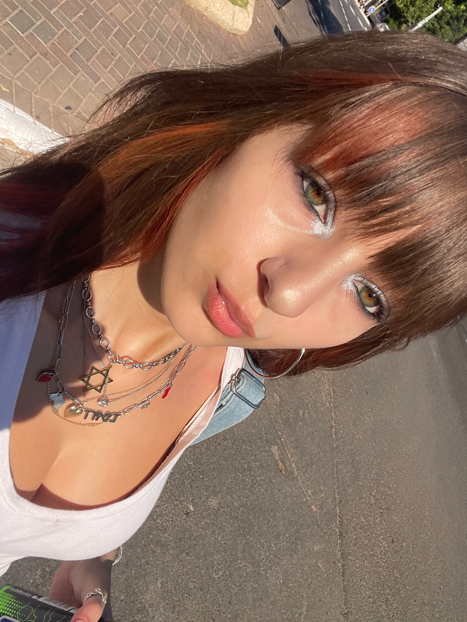 A young woman with brown hair with red highlights, green eyes, and full lips is shown in a close-up, outdoor portrait. She is wearing a white tank top and multiple necklaces, including a Star of David pendant. The background shows a brick sidewalk and a street, suggesting an urban environment. The image appears to have been taken in Raanana, Israel, as indicated by the context. The lighting suggests it is daytime, with sunlight casting shadows.