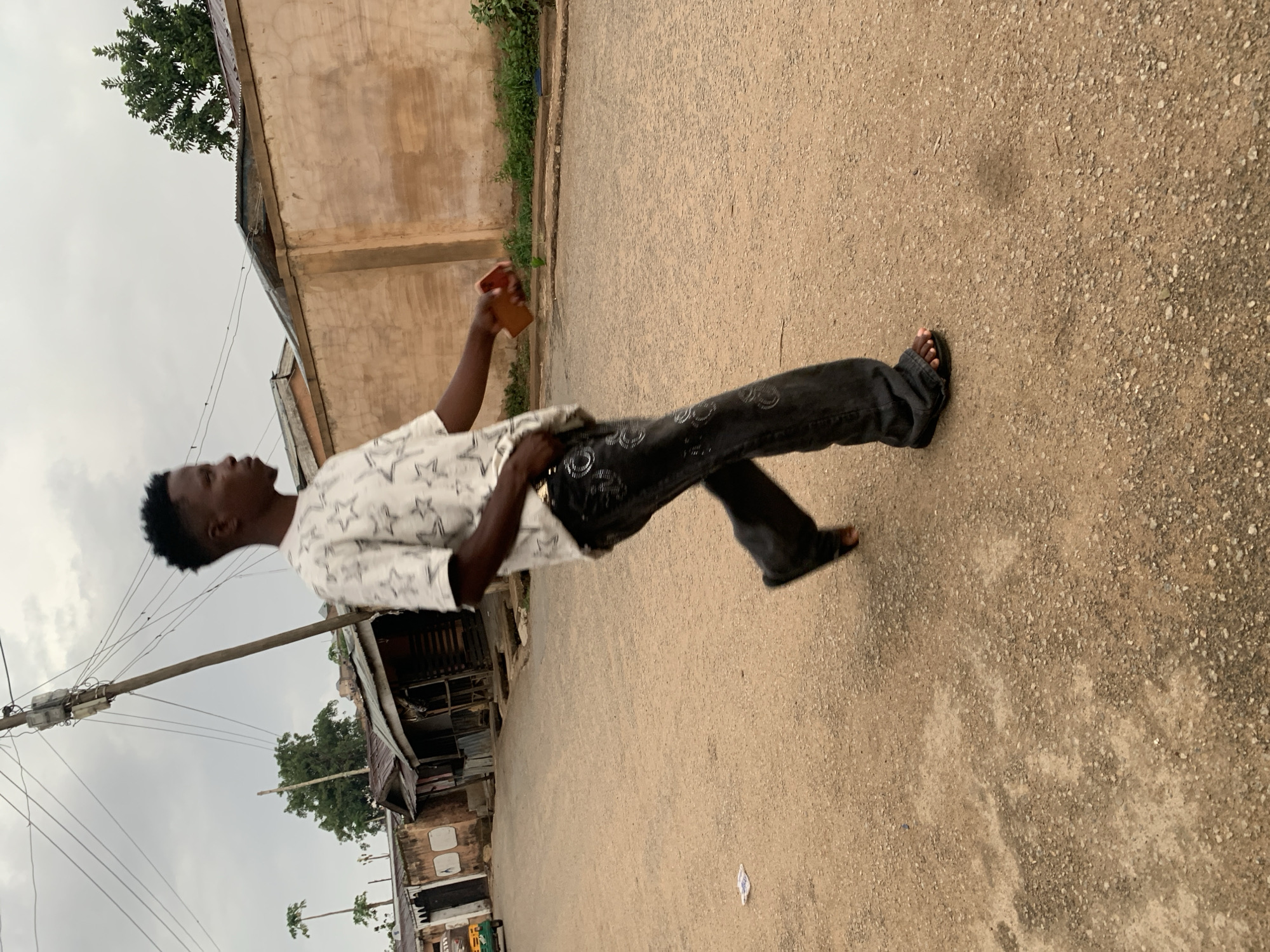 The image depicts a young Black man walking on a paved road in Jos, Nigeria. He is holding a smartphone in his right hand and appears to be looking at it. He is wearing a white star-patterned shirt and black jeans. He is wearing black sandals. The road is bordered by buildings, some of which appear to be residences or shops. Overhead wires are visible. The sky is overcast. The terrain consists of light-colored dirt.