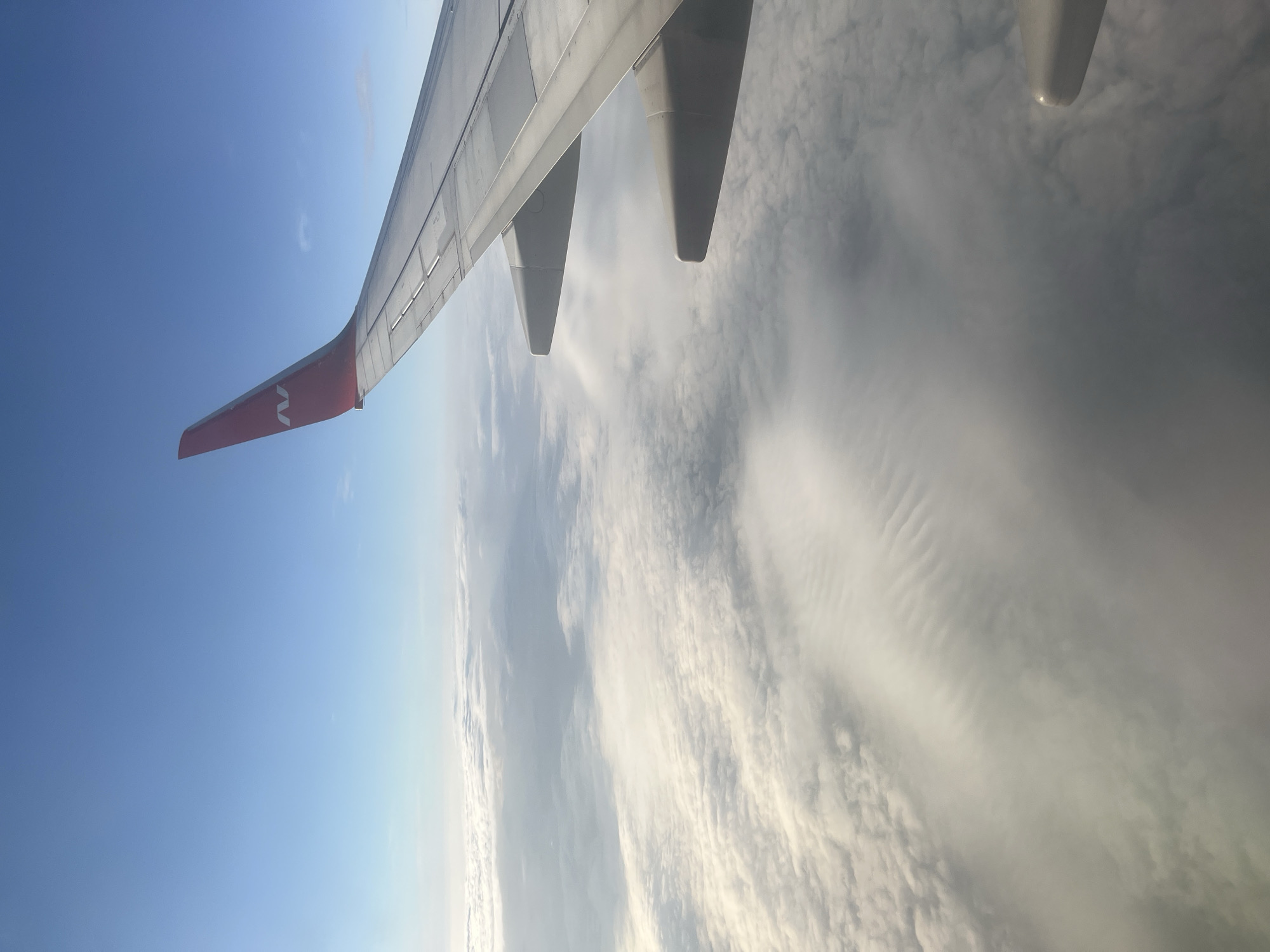 An aerial view captures the wing of a passenger airplane against a backdrop of a clear blue sky and a vast expanse of clouds. The wing, painted white with a red tip bearing a stylized "N" logo, extends diagonally across the right side of the frame. Below the wing, a thick layer of white clouds fills the lower portion of the image, creating a textured, undulating landscape. The clouds are illuminated by sunlight, casting shadows and highlighting their forms. The scene is static and peaceful, conveying a sense of travel and the beauty of the atmosphere from above. The location is identified as Stolbishche, Russia.