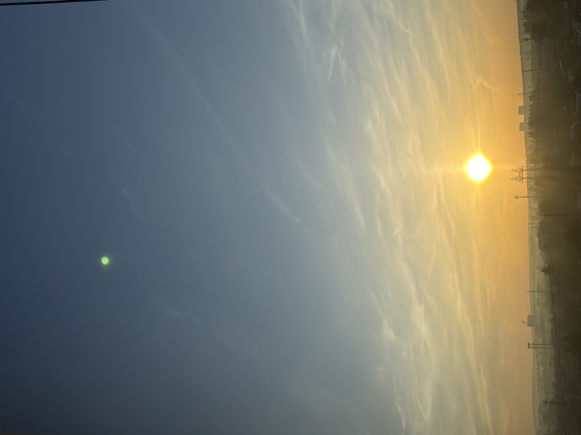 The image shows a view of Chaikovsky, Russia at sunrise or sunset. The sun is visible as a bright disk on the right side, near a cityscape silhouette including buildings and power lines. The sky is divided into a gradient, transitioning from a blue on the left to yellow and orange on the right where the sun is positioned. Wispy clouds are present in the sky, and there's a green dot visible in the blue portion, which might be a lens flare.