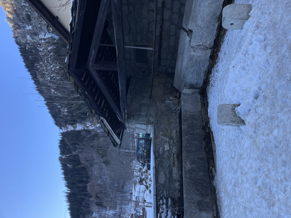 This image captures a serene winter landscape in a mountainous region, consistent with the Courmayeur, Italy, context. The scene is dominated by a traditional stone building with a dark wooden roof structure, visible on the right side of the frame. Its sturdy stone walls and exposed timber beams suggest classic alpine architecture.
The foreground and mid-ground are covered in a blanket of snow, with some patches of bare earth visible. Two distinctive, rectangular stone blocks, possibly bollards or markers, are partially submerged in the snow close to the viewer. A longer, lower stone structure runs parallel to the building's base, forming what appears to be a pathway or a low retaining wall.
Further back, near the building, a dark-colored vehicle, resembling an excavator or small tractor, is parked on the snowy ground. Beyond the building and vehicle, a rugged mountain slope rises majestically, densely covered with a mix of bare deciduous trees and dark green coniferous trees, indicating a forest. The upper portion of the scene is dominated by a clear, vibrant blue sky.
The time of day appears to be daytime, with strong, clear light suggesting a sunny winter day. There is no visible activity taking place, contributing to a peaceful and quiet atmosphere. No people are visible, and there is no discernible text in the image. The overall impression is one of tranquil natural beauty combined with traditional mountain habitation.
