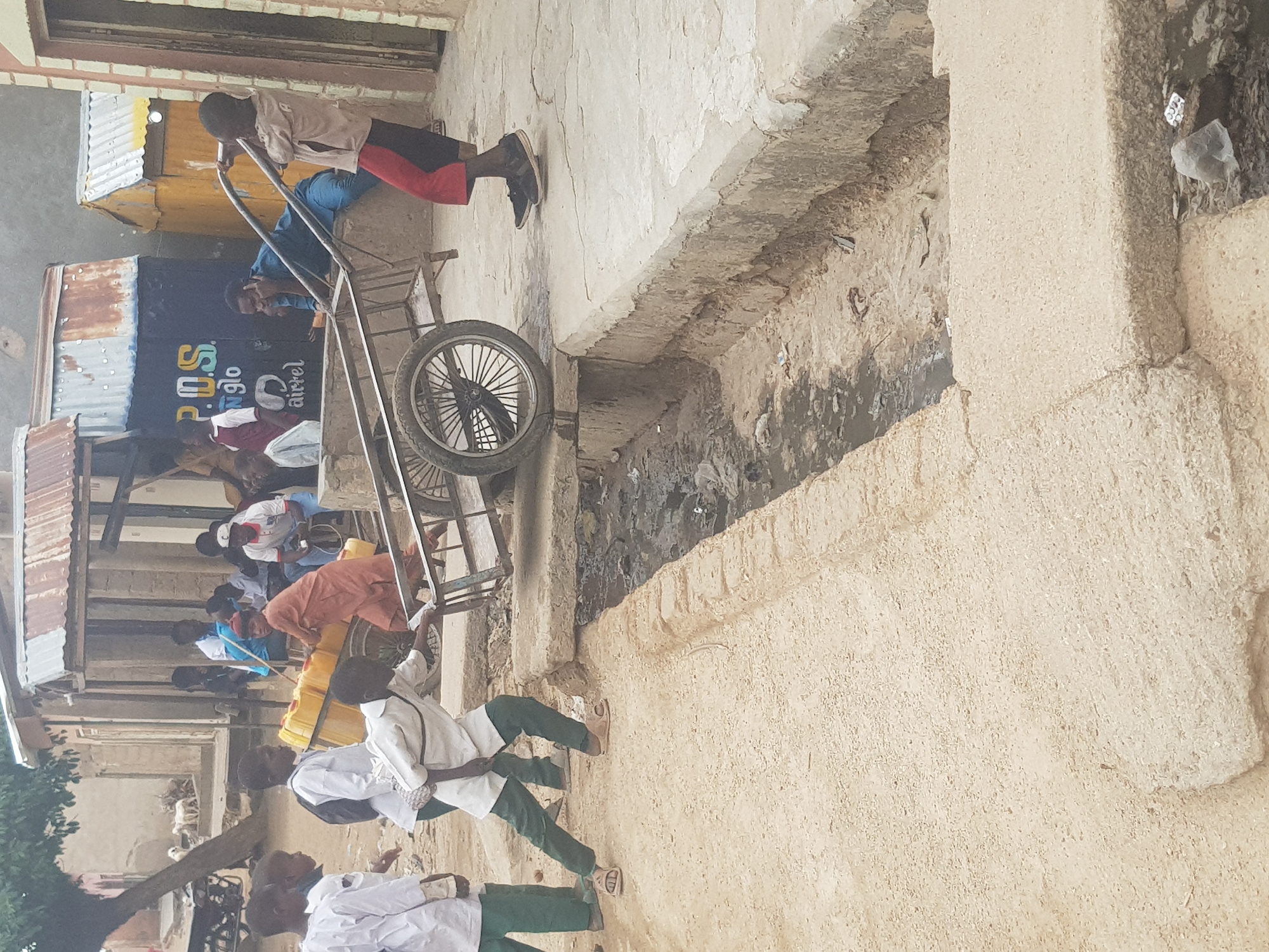 The image depicts a street scene in Dabawa, Nigeria. A group of people are gathered near a P.O.S. (Point of Sale) store. Several individuals are transporting yellow jerrycans, possibly containing water, using a metal cart. The street is narrow, with concrete structures and a drainage ditch visible. Some people are wearing white shirts and green pants, which might be school uniforms.