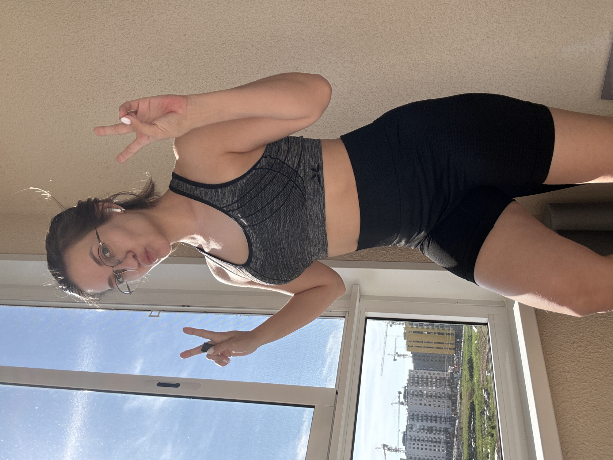 A young woman is captured in a full-body, portrait-style photograph, presented as if rotated 90 degrees counter-clockwise. She stands indoors near a large window, dressed in athletic wear: a grey speckled sports bra with subtle black patterned lines and a stylized 'X' logo, paired with form-fitting black shorts. Her dark hair is pulled back, and she wears round glasses. She makes a playful puckering face towards the camera and forms a "peace" sign with both hands, with a dark ring visible on her right index finger and white nail polish. An earbud appears to be in her left ear.
The indoor setting features a textured, light-colored ceiling or wall. Through the expansive, white-framed window, a bright, clear blue sky is visible in the upper portion. The lower part of the window reveals an urban landscape, indicative of Pervomaysky, Russia, under active development. Several large, modern apartment buildings are visible, some with prominent construction cranes towering beside them. Patches of green land stretch between the buildings. The bright natural light indicates it is a clear, sunny daytime. The overall mood of the image is casual and lighthearted. No explicit text is visible beyond the small logo on her sports bra.