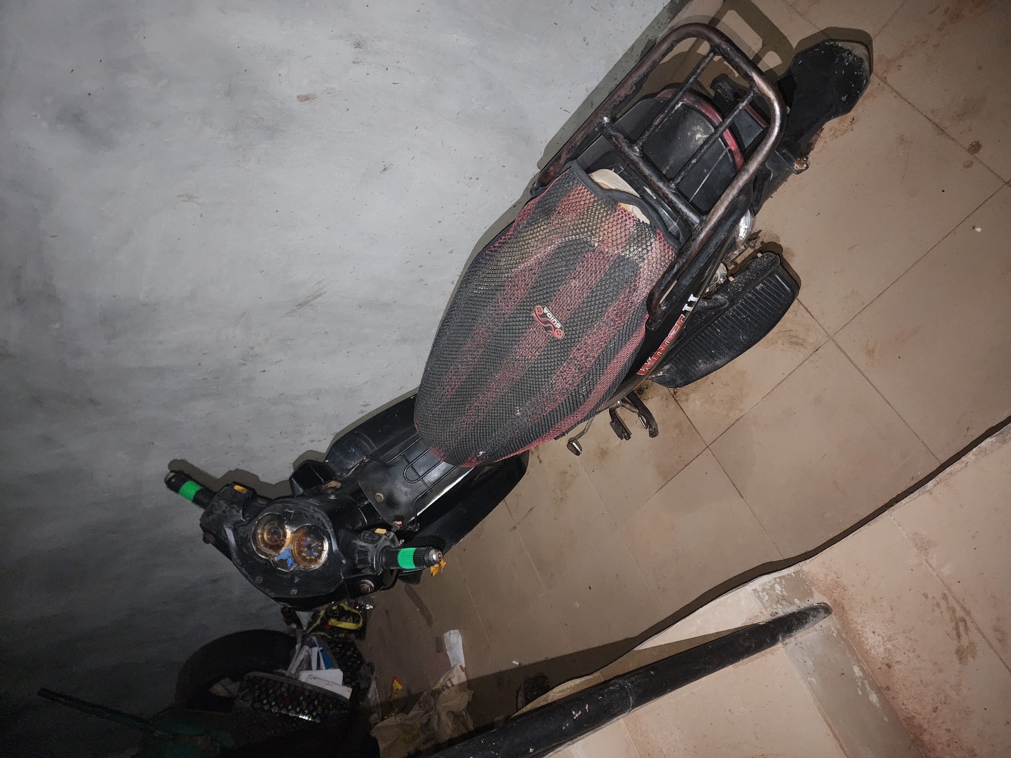 The image shows a motorcycle parked indoors. The location is likely Abraka, Nigeria, based on the prompt. The motorcycle has a black seat cover with red accents, green handlebars, and a luggage rack. The floor is tiled, and the walls are painted. There is no information in the image about people, actions, or interactions.