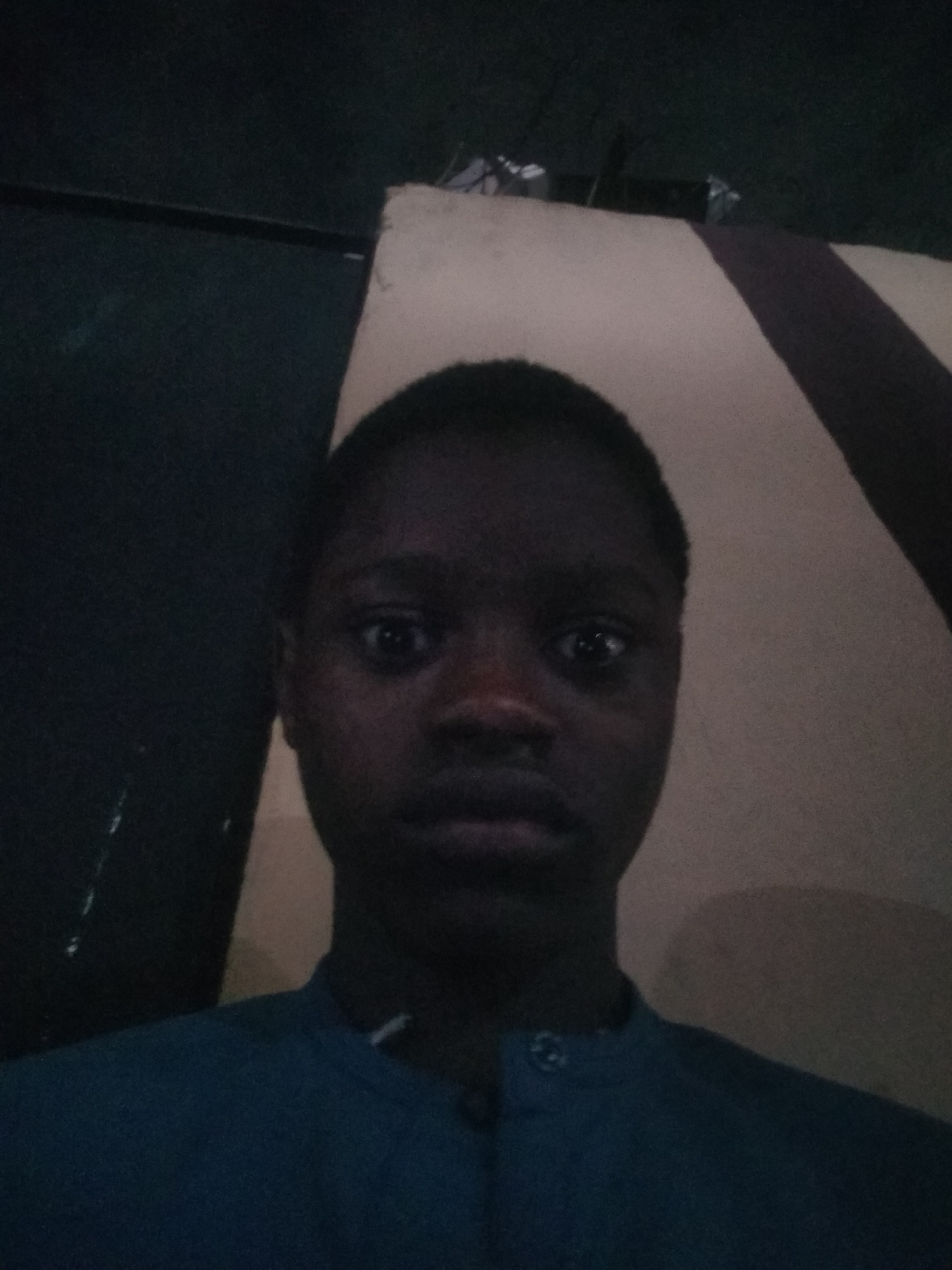 A medium close-up selfie shot features a dark-skinned person with short, dark hair. The individual is wearing a button-down shirt. The person's face is centered and fills much of the frame, drawing attention to their eyes and features. The background consists of two distinct areas: a dark, undifferentiated area on the left and a lighter area with dark brown stripes on the right. This suggests the person is indoors. The image doesn't allow for a confirmed description of Shangisha, Nigeria.