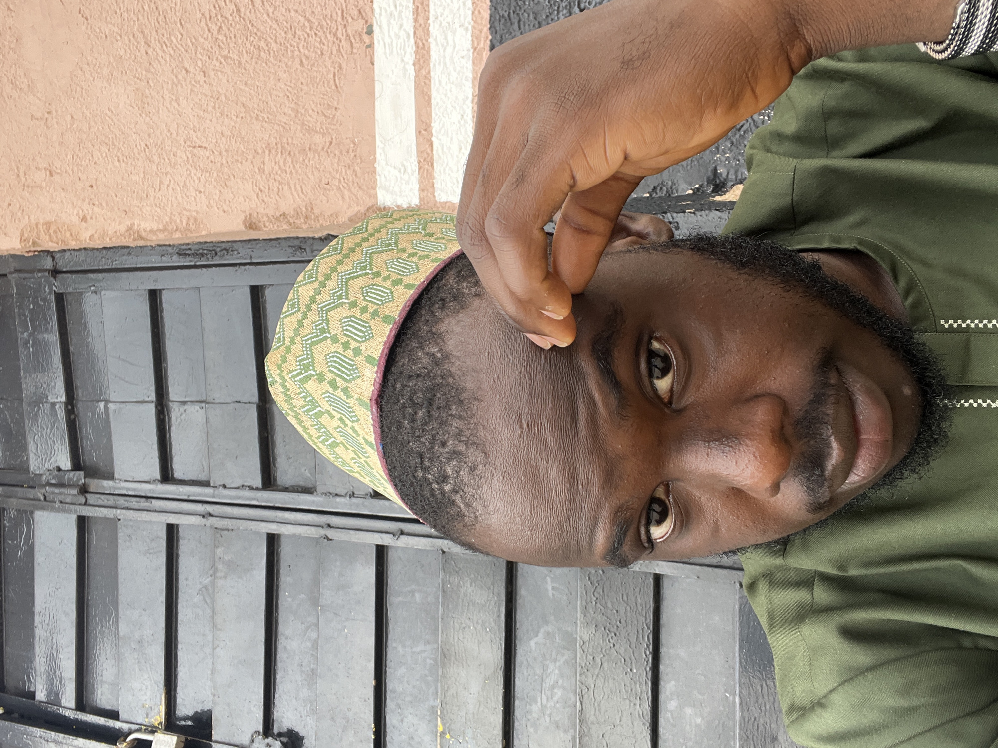 The image presents a close-up shot of a dark-skinned man, possibly in his late 20s to early 30s. He is wearing a green traditional hat with a geometric pattern and a matching green shirt. The man's right hand is raised, with his fingers touching his forehead.
The background is a combination of textures and colors. Behind the man's head is a gray, metal gate-like structure. Adjacent to the gate is a light brown wall with a textured surface. In the upper part of the image, there's a section with white stripes.
Based on the man's attire, the presence of the traditional hat, and the information provided, it is likely that the image was taken in Jos, Nigeria.