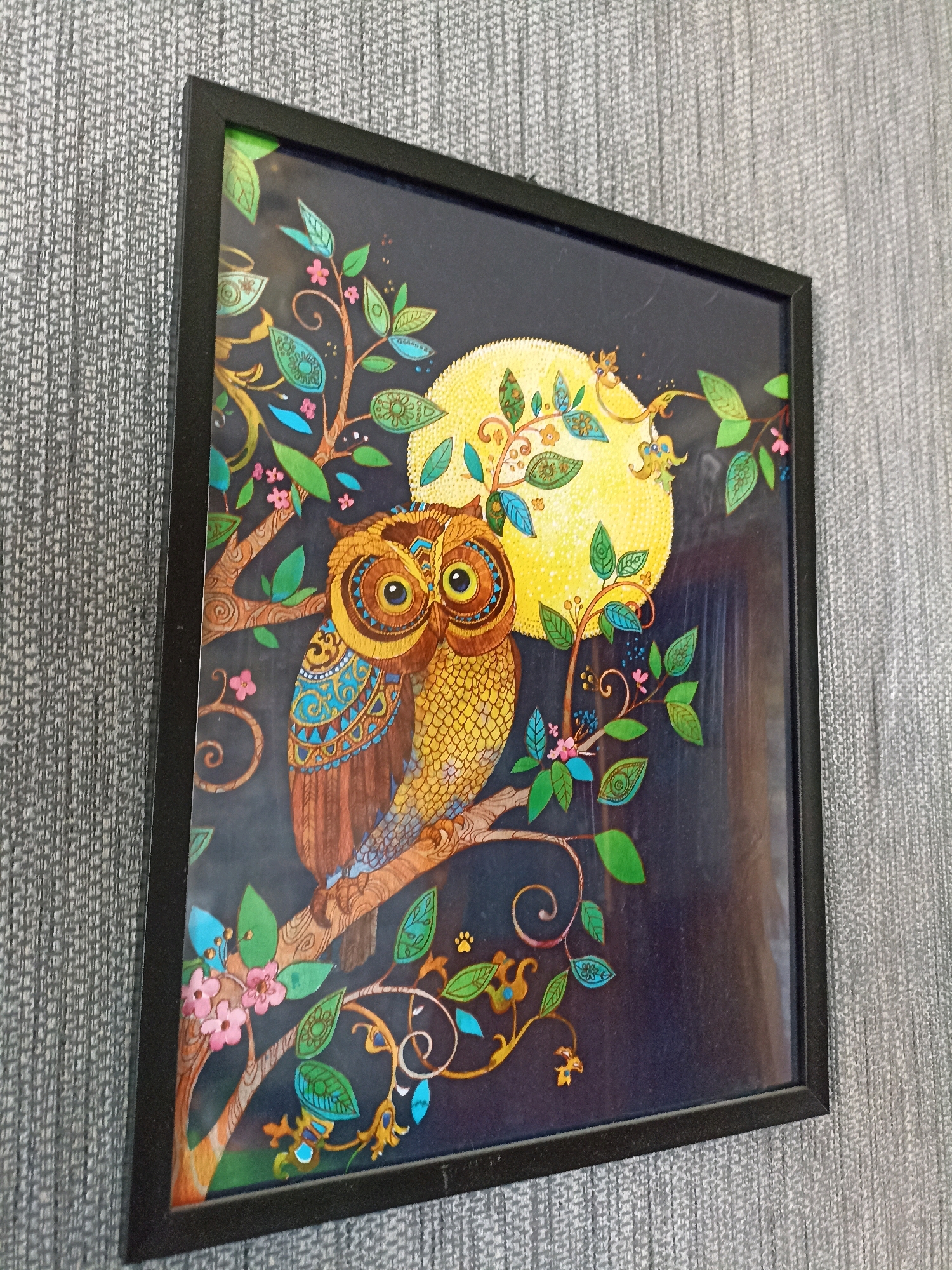 A framed picture of two owls perched on a branch against a dark background is hanging on a textured gray wall. The picture depicts a stylized scene with a large yellow moon, colorful foliage, and intricate patterns on the owls' feathers. The frame is black and rectangular. The scene is illuminated by ambient light, casting faint reflections on the glass of the frame. There are no people or animals present in the image other than those depicted in the artwork. The location is identified as Novosibirsk, Russia.