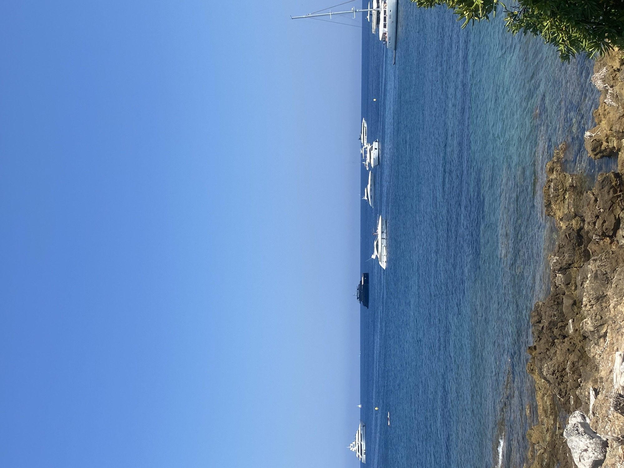 The image depicts a serene seascape with several yachts and sailboats anchored in the blue waters off the coast of Antibes, France. In the foreground, rugged, rocky terrain meets the clear, tranquil sea, with a hint of green foliage on the right. The sky above is a vast expanse of clear, bright blue. Several yachts of varying sizes and designs are visible on the water, along with a few sailboats. One large white sailboat with a tall mast is prominently positioned on the right side of the frame. The water is calm, with gentle ripples and some areas of transparency revealing the seabed. The overall scene suggests a leisurely day at sea, with the yachts appearing to be at anchor.