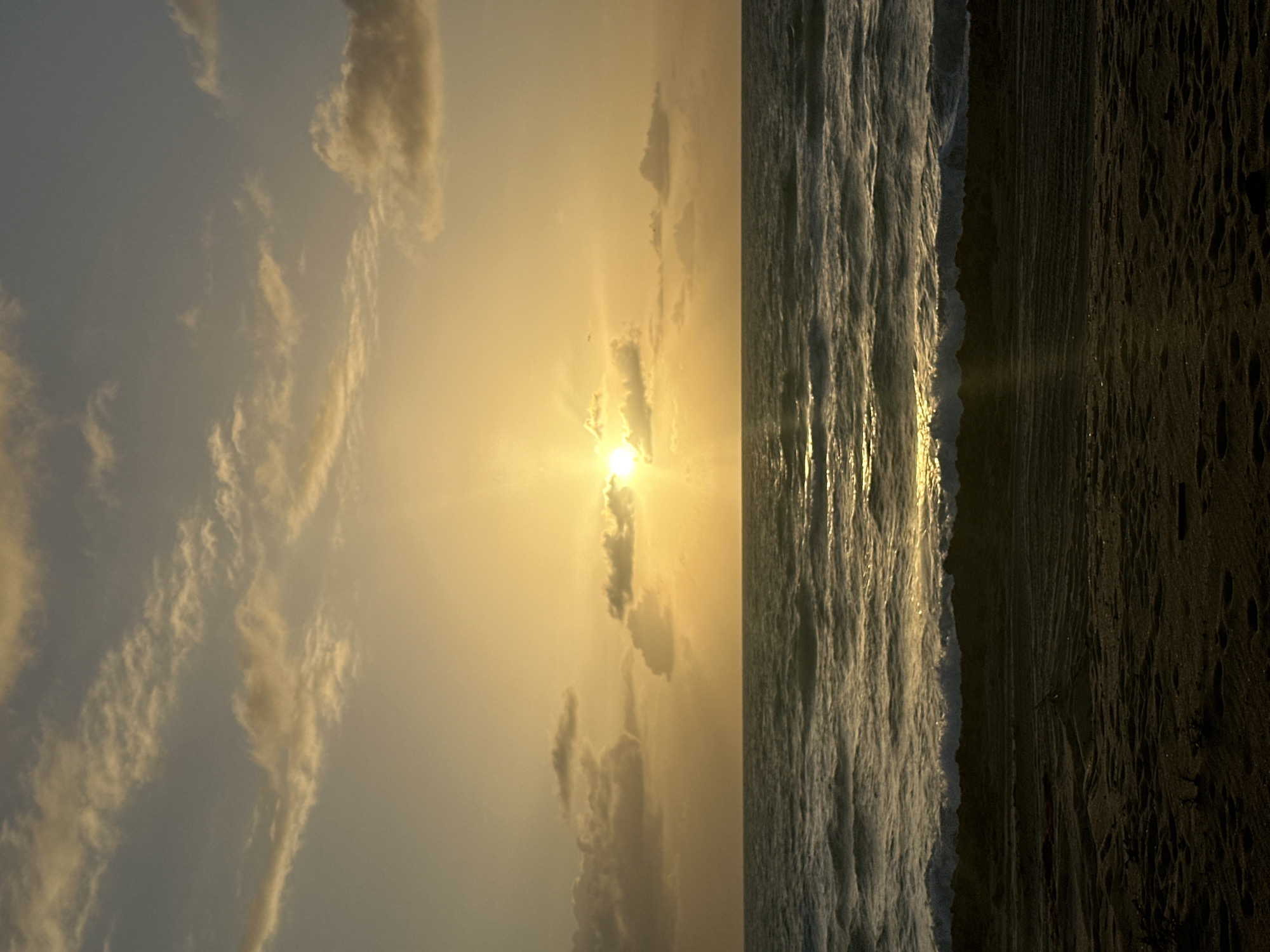 The image captures a sunset scene at Hallandale Beach, Florida, United States. The sky is filled with clouds, illuminated by the golden light of the setting sun. The ocean waves crash onto the dark, sandy beach, reflecting the sunlight. No people are visible in the scene.