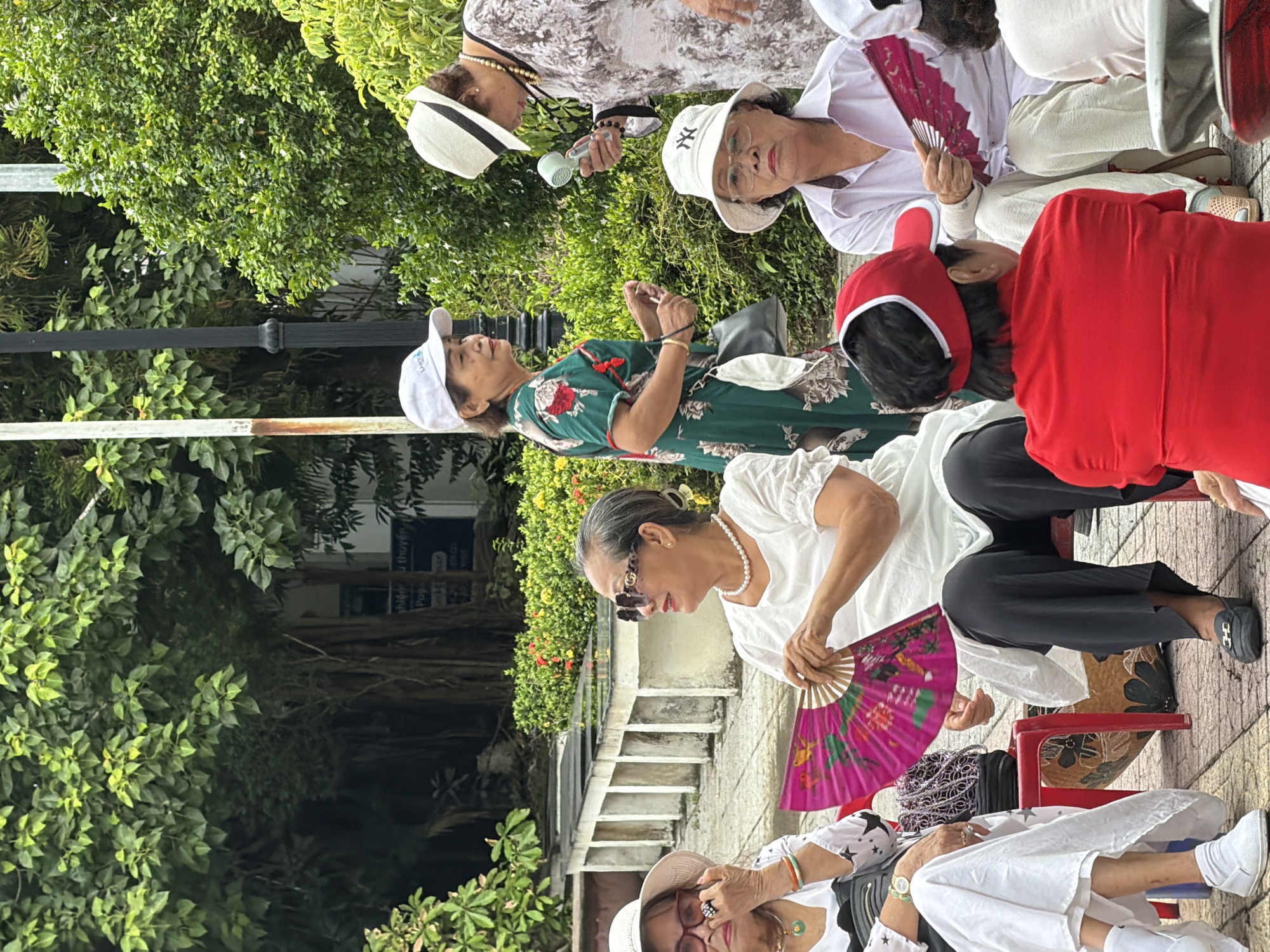 In Thuận Hóa Ward, Vietnam, a group of elderly individuals are gathered outdoors, seemingly in a public park or plaza. Several women are seated on red stools and chairs. One woman, wearing a white top and black pants, is fanning herself with a pink oriental fan. Another woman in a white outfit is partially visible on the left, her face obscured by a hat and her hand touching her face. A third woman, in a green floral dress and white cap, stands holding a small, handheld fan. To her right, a woman in a patterned dress and white hat appears to be speaking to someone off-camera, holding a small, light blue fan. A woman in a white shirt and hat sits in the foreground, looking forward. The ground is paved with cobblestones. Lush green trees and bushes form the background, with some architectural elements like low concrete walls and planters visible. The lighting suggests it is daytime.