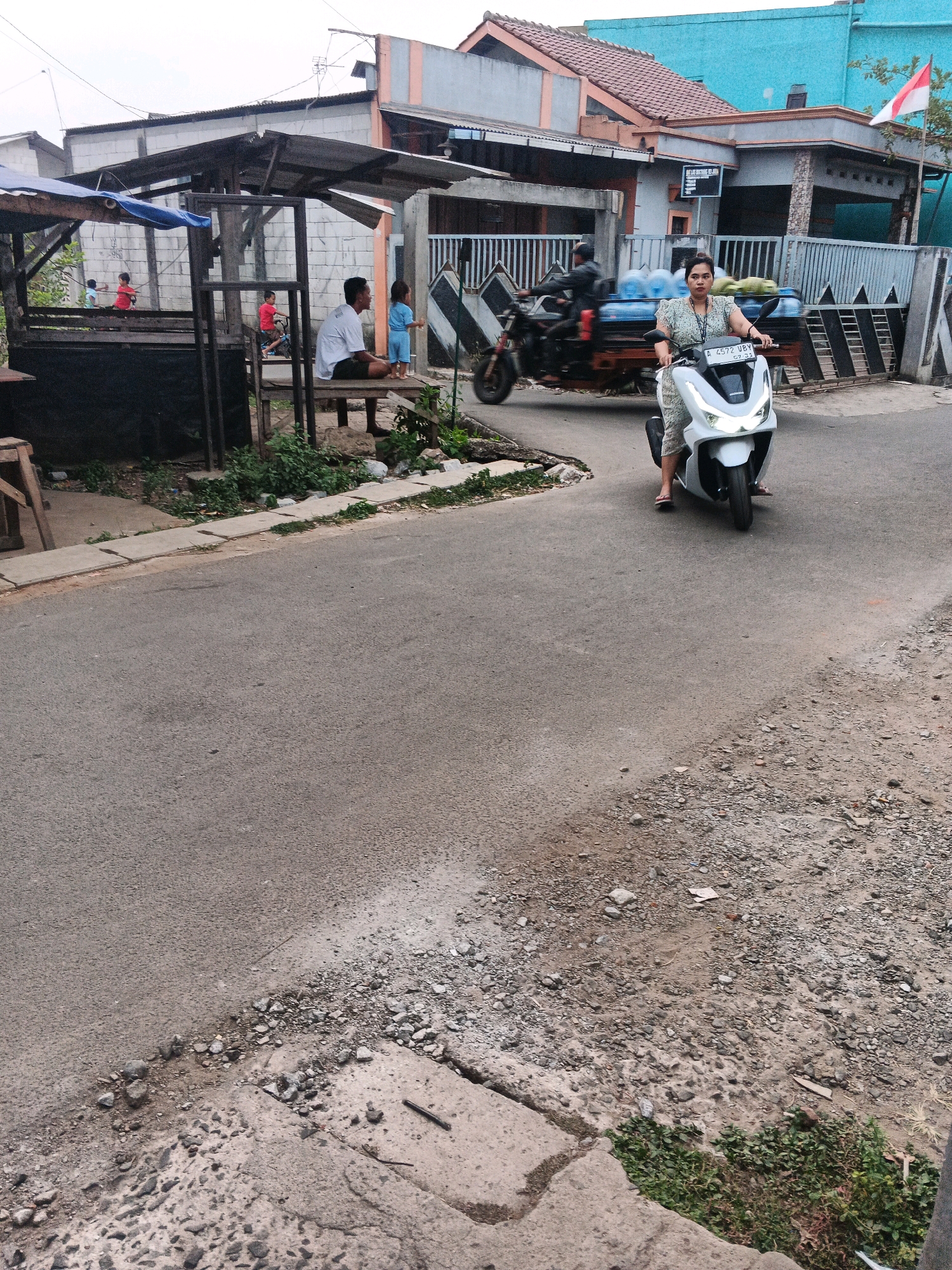 This is a photograph taken in the city of Kadu, Indonesia. The scene depicts a street with several elements of daily life. In the foreground, a woman is riding a white Honda motorcycle, looking directly at the camera. She is wearing a light-colored patterned dress and flip-flops. Behind her, a man is riding a motorcycle with a trailer loaded with water gallons and coconuts. In the background, there is a covered wooden structure with people, including children, some sitting and others standing. A man in a white shirt and dark shorts is seated, and a child in blue shorts stands beside him. The buildings in the background are a mix of residential structures, one with a blue facade and a red-tiled roof, and another with a gray and orange color scheme. A small Indonesian flag is visible on a pole in front of one of the houses. The street surface is asphalt with scattered gravel and some concrete elements. The overall atmosphere suggests a typical day in a semi-urban or residential area.