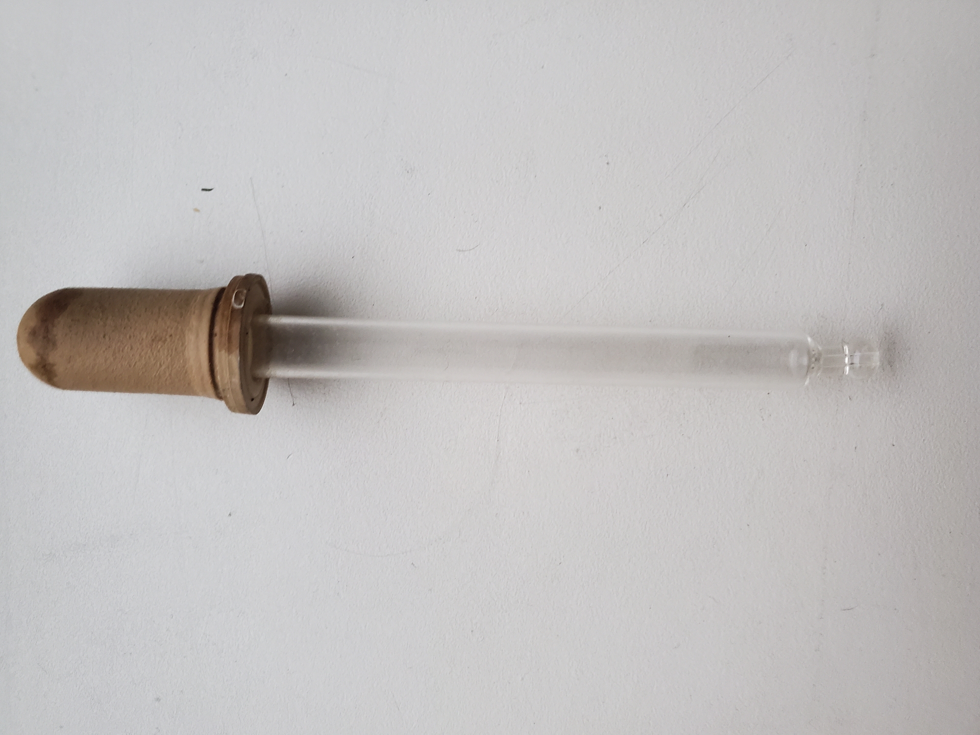 A glass dropper with a brown rubber bulb is positioned vertically on a textured white surface. The dropper has a clear glass tube with a bulbous tip at the bottom. The rubber bulb at the top is worn and a light brown color. The surface it rests on has faint lines and specks, suggesting it is a wall or similar textured material. The image is taken from a top-down perspective. There is no indication of the city of Tiraspol, Moldova, or any other location.