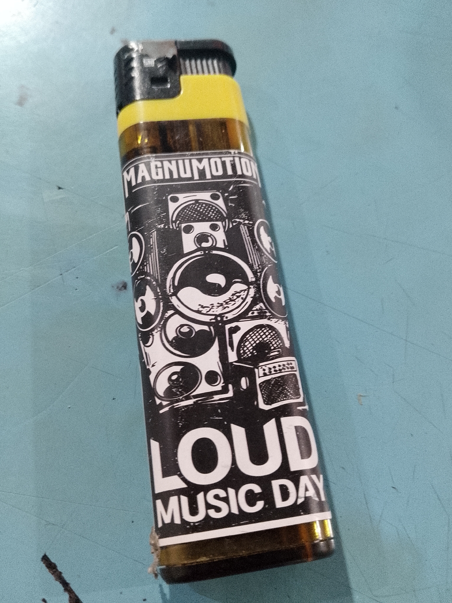 A close-up shot shows a lighter resting on a light blue surface. The lighter has a yellow top and a transparent body, allowing a view of the fuel level inside. A black and white design wraps around the body, featuring illustrations of speakers and amplifiers in a graffiti style. The words "MAGNUMOTION," "LOUD," and "MUSIC DAY" are printed in bold, capital letters. The lighter's black flint wheel and gas nozzle are visible. The image was probably taken indoors, and it's impossible to pinpoint the location in Lhokseumawe, Indonesia from the photo.