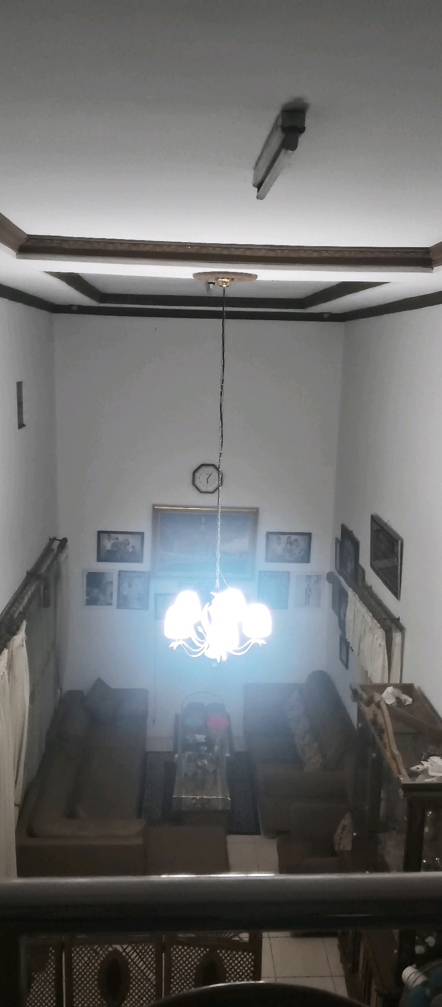 The image depicts an interior view of a room, likely a living area, with a high ceiling. A prominent chandelier with multiple bulbs illuminates the space, casting a bright glow. Several framed pictures are hung on the white walls. In the background, sofas and a coffee table are visible, suggesting a seating arrangement. The overall impression is of a domestic environment. Based on visual cues and general knowledge of common interior designs in residential areas, this scene is consistent with a home located in Cianjur, Indonesia.