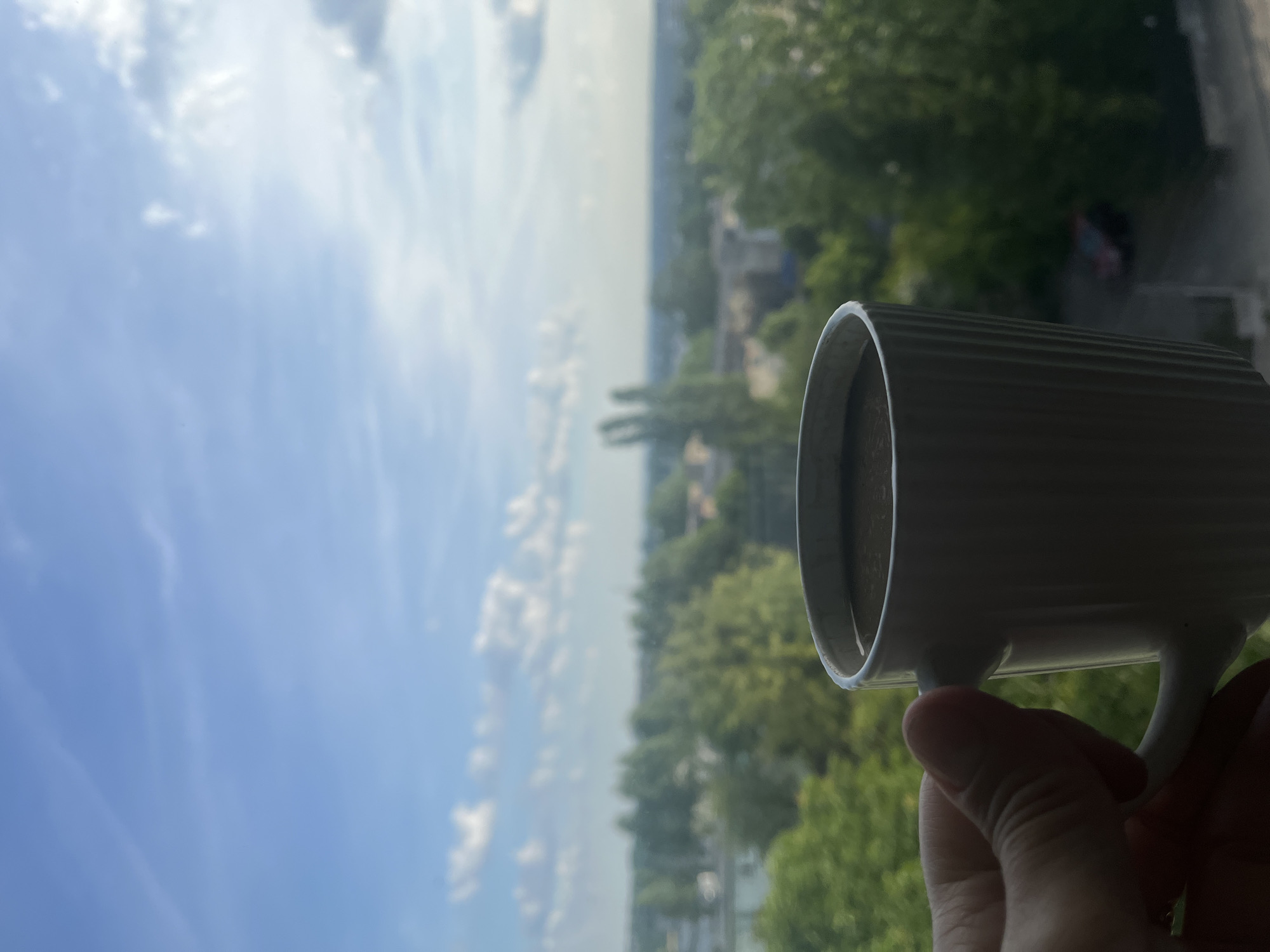 A hand holds a white mug of dark liquid, likely coffee, in front of a blurred background of a city skyline. The sky above is blue with scattered white clouds. Trees and buildings are visible in the mid-ground, suggesting an urban or suburban setting. The lighting indicates it is daytime, possibly morning or late afternoon.