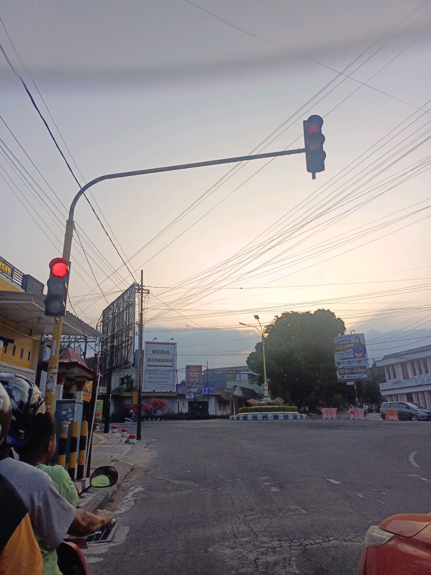 This is a photograph taken at a street intersection in Candirejo, Indonesia, at dusk. The scene is dominated by a traffic light displaying red, positioned above a streetlamp and an array of overhead electrical wires that crisscross the sky. In the foreground, the back of a motorcycle with two passengers is visible, suggesting movement or a pause at the intersection. To the left, a building with yellow accents lines the street, and to the right, a roundabout with a decorative planter is present. Billboards, one advertising "MEDIA DISEWAKAN" and another featuring an advertisement for "KREDIT MULTIGUNA," are prominent features of the urban landscape. A dark-colored car is seen driving away in the background. The sky is hazy with the soft light of twilight.
