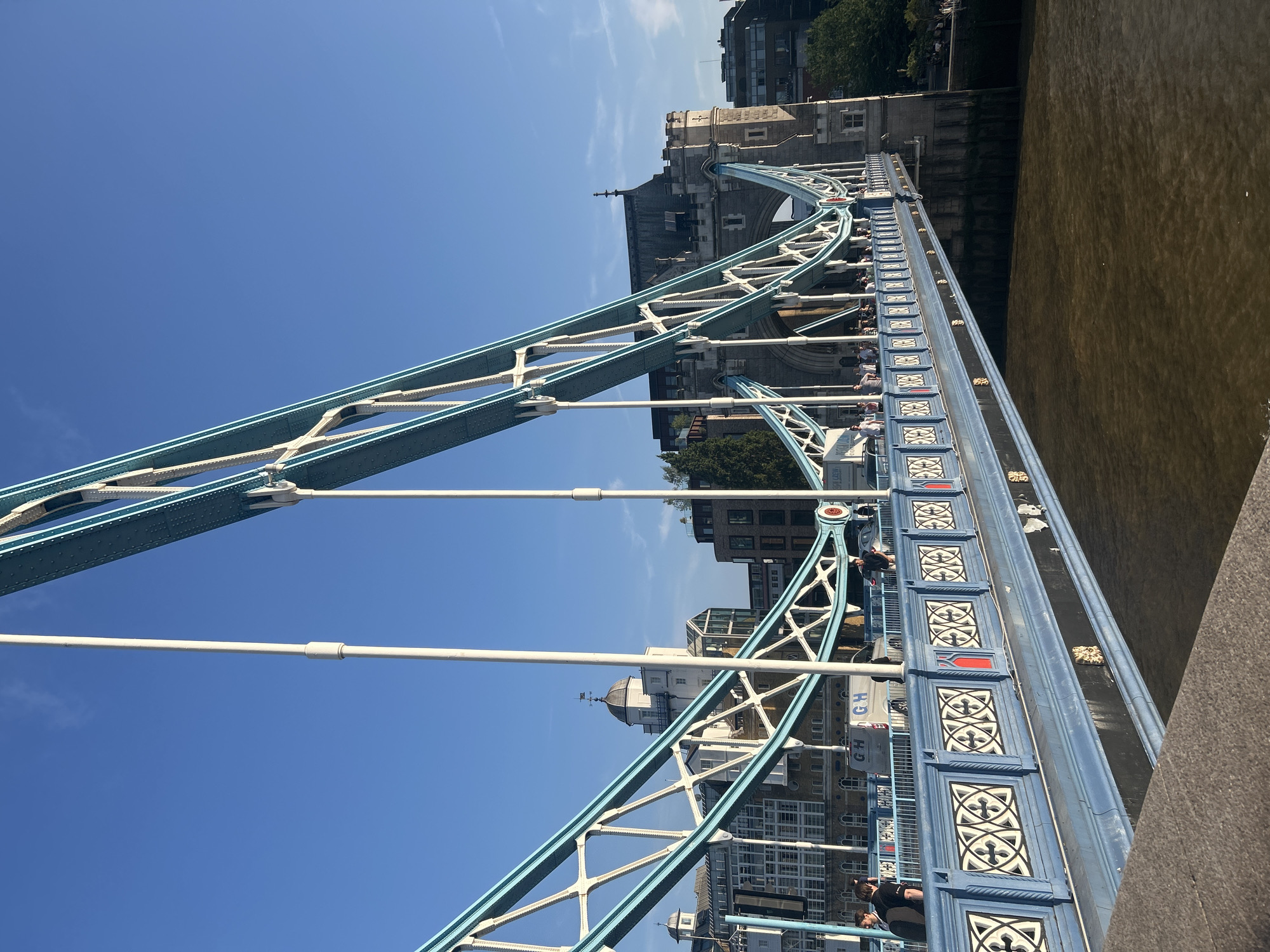 This is a low-angle, full shot of Tower Bridge in London, United Kingdom, under a clear blue sky. The bridge's distinctive blue and white structure dominates the foreground, with its intricate lattice work and suspension cables stretching towards the top of the frame. A pedestrian walkway is visible on the bridge, lined with a decorative blue railing featuring white and red patterns. Several people are walking along the bridge, some appearing to be tourists. In the background, the iconic stone towers of Tower Bridge are visible, along with several buildings on the north bank of the River Thames. The water of the Thames, a murky brown, flows beneath the bridge. The lighting suggests it is either early morning or late afternoon, with the sun casting long shadows.
