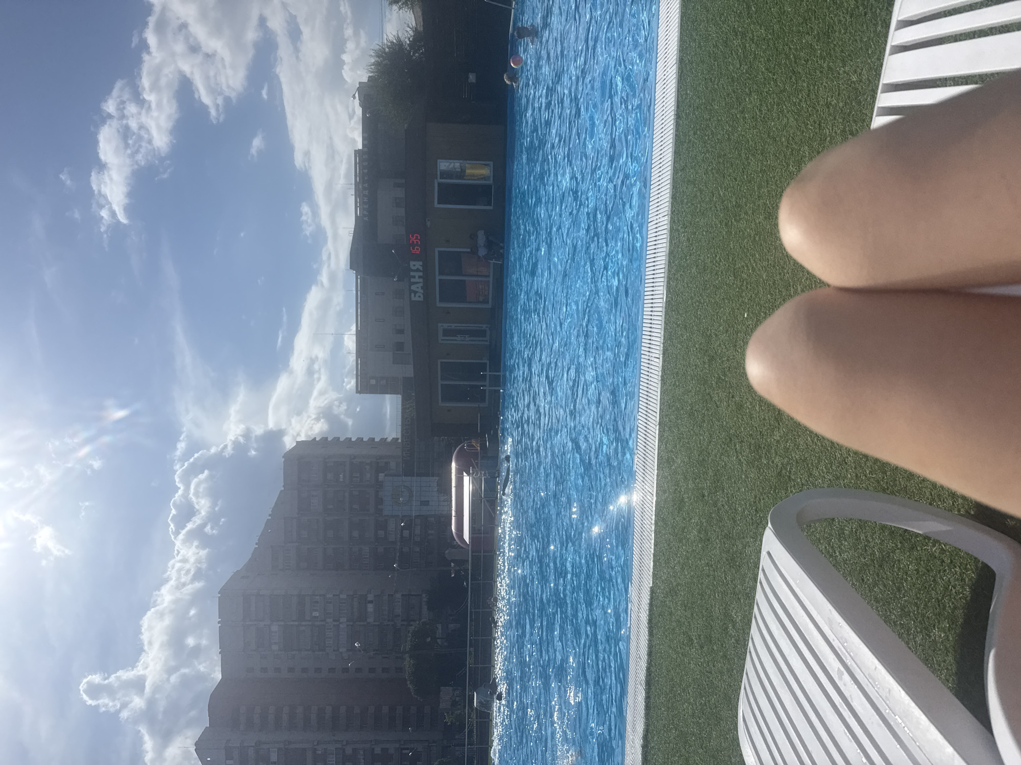 The image captures a sunny day at an outdoor swimming pool, likely in Magnitogorsk, Russia, given the signage. In the foreground, the legs of a person are visible, resting on a white lounge chair with a ribbed texture, positioned on a patch of artificial turf.
Beyond the lounge chair, a large, bright blue swimming pool dominates the midground. The water is clear and reflects the sunlight, creating shimmering patterns. Several people are present in the pool: some are swimming, one individual is playing with a pink ball, and another appears to be partially submerged. In the background, a building with "БАНЯ" (Banya, meaning bathhouse in Russian) displayed in red digital numbers shows "16:35". Adjacent to this building, other structures are visible, including taller apartment buildings that appear to be residential complexes, suggesting an urban setting. The sky is blue with scattered white clouds and the sun is shining brightly, casting shadows. A red inflatable structure is partially visible within the pool area.