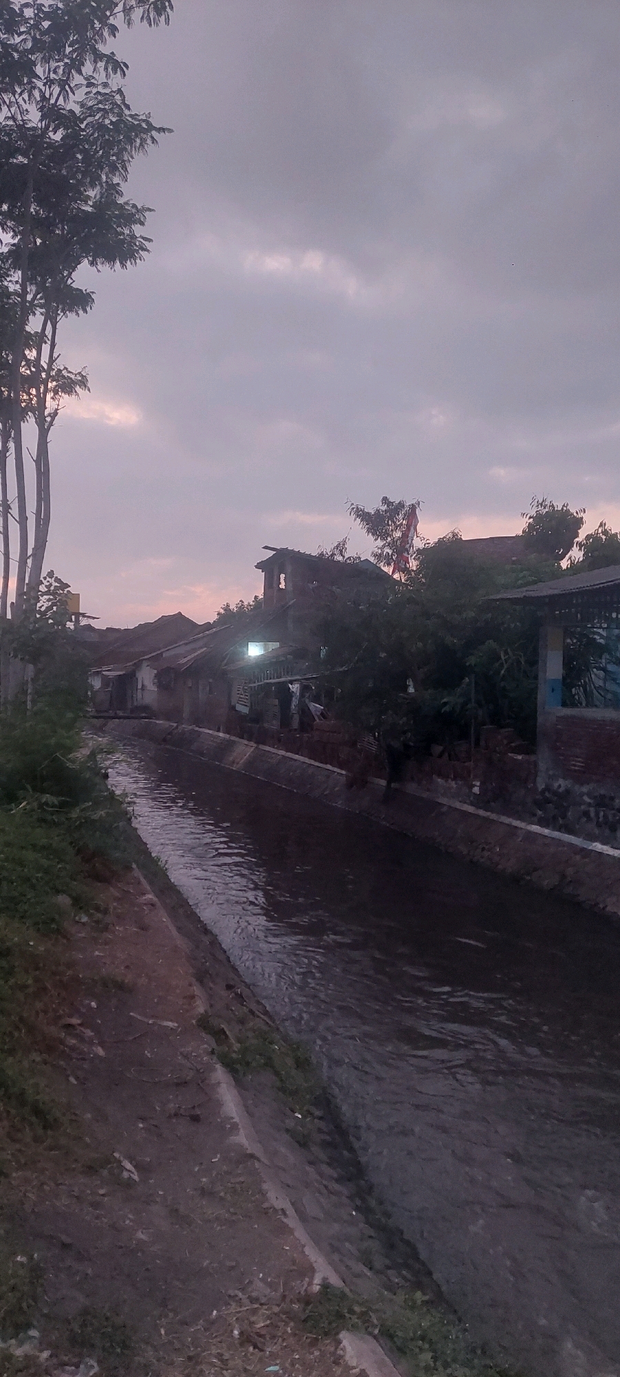 In Jogoyudan, Indonesia, a village scene unfolds during twilight, with a narrow canal flowing through its center. The canal, lined with concrete and rough earth, reflects the dimming sky and the silhouettes of houses on its banks. Tall, slender trees stand on the left, their foliage dark against the muted purple and grey clouds. The houses, constructed with various materials including corrugated metal and brick, show signs of wear and tear. Some have visible light sources, suggesting ongoing activity. A red and white flag, possibly the Indonesian flag, is partially visible on a pole behind the houses. The overall atmosphere is calm and still, characteristic of a rural Indonesian village at dusk.
