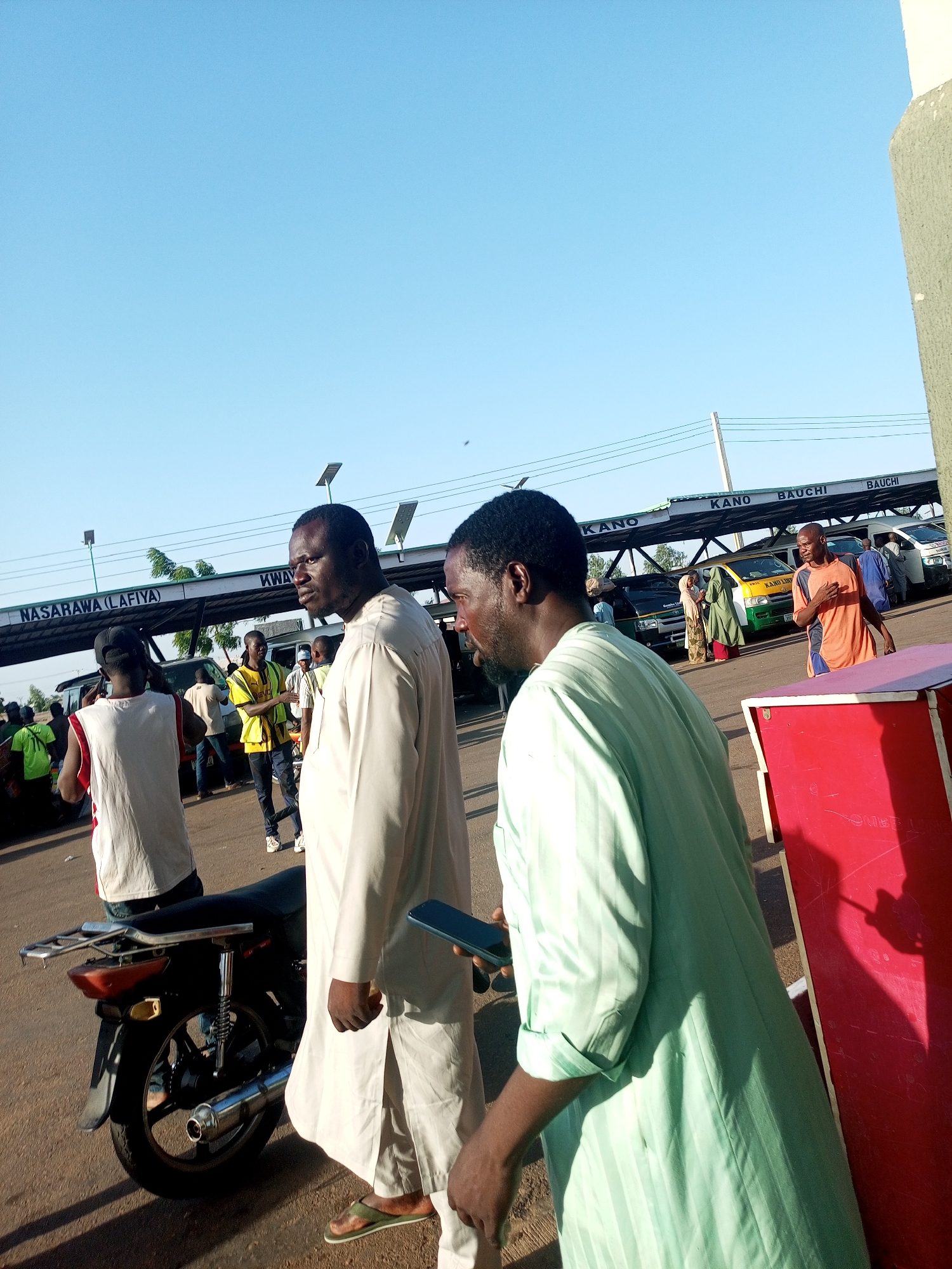 The image shows a bustling outdoor scene in what appears to be a bus or transportation terminal, with multiple people and vehicles present. In the foreground, two men are walking. The man on the right is wearing a light green, striped traditional garment and holding a smartphone. The man to his left is wearing a light-colored, likely white, traditional gown with matching trousers, with brown sandals.
To the left, a man in a white tank top sits on a motorcycle. Behind the men, there are other individuals and commercial buses, painted in various colors and marked with destinations like "KANO" and "BAUCHI." The overhead structure is a signpost indicating destinations, including "NASARAWA (LAFIYA)." Solar panels are mounted on the structure.
The environment indicates a sunny day with a clear blue sky. The ground is paved, and there is some debris scattered on the road. In the background, there is more activity with individuals standing near the buses and people moving about, suggesting a busy hub of transportation. There is a red rectangular object on the right side of the image. The city is unknown.