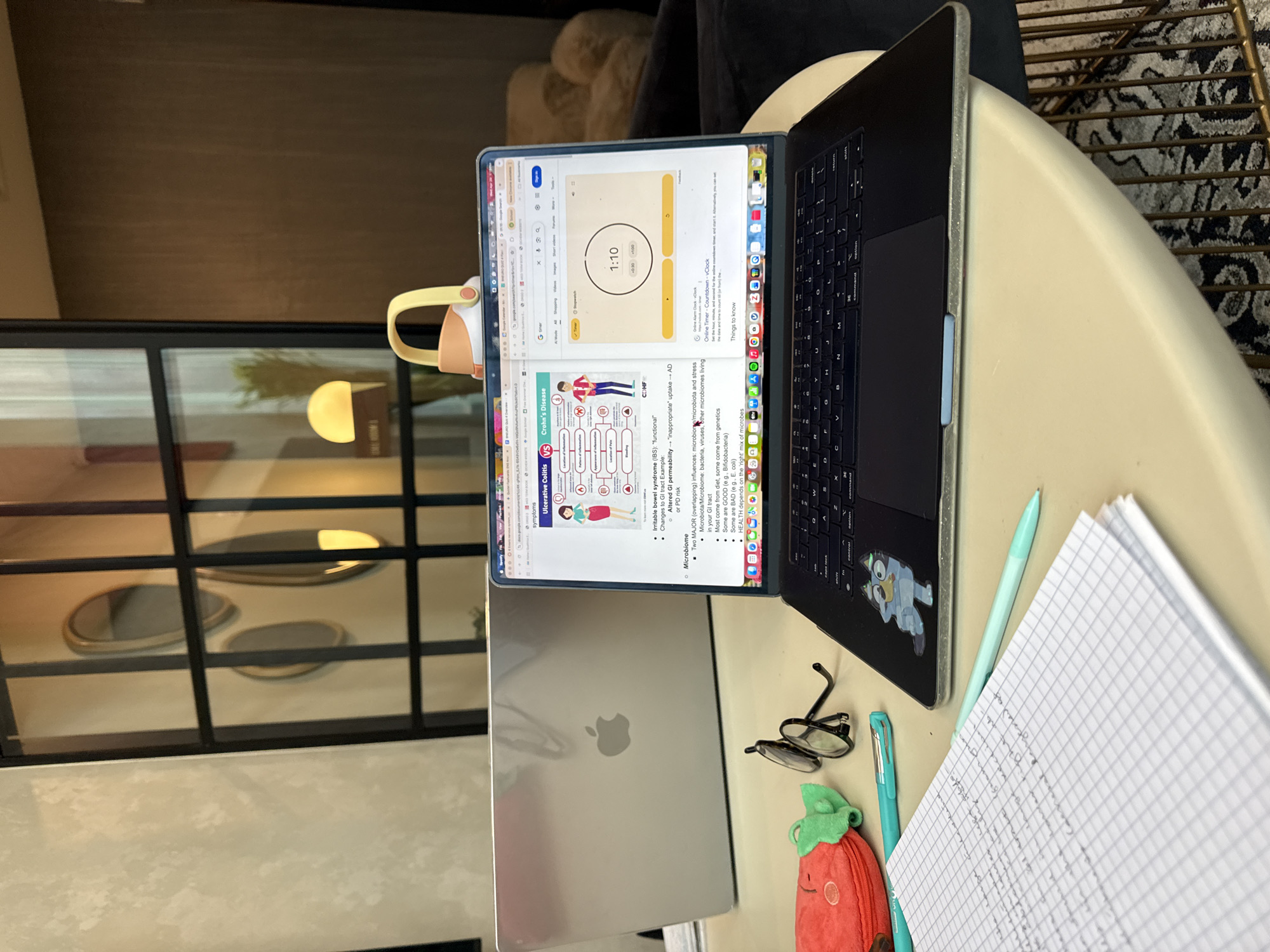 An overhead shot captures a workspace setup on a light-colored table. The main subject is an open Apple MacBook Pro laptop, positioned with its screen facing slightly left. The screen displays a web browser, prominently featuring a "G timer" set to "1:10" and notes related to health and medical topics. A diagram titled "Ulcerative Colitis VS Crohn's Disease" is visible, along with text sections on "Microbiome," "Irritable bowel syndrome (IBS)," and "Altered GI permeability." Other visible text includes "Online Alarm Clock - vClock," "Online Timer - Countdown - vClock," and bullet points detailing "Two MAJOR (overlapping) influences: microbiome/microbiota and stress," "bacteria, viruses, other microbiomes living in your GI tract," and "HEALTH depends on the 'right' mix of microbes."
To the left of the open laptop, a closed silver Apple MacBook reflects the screen of the active laptop. A yellow-handled water bottle is partially visible next to the closed laptop. On the right side of the active laptop's keyboard, a light blue cartoon sticker of a character, likely from "Bluey," is affixed. Beside the laptop are a pair of black eyeglasses, a light blue pen, and a teal pen. A red, strawberry-shaped plush item or pouch lies near the glasses. Further right, a grid-lined notebook with illegible handwriting is open.
The background shows an indoor setting, likely a modern room. Behind the table, a textured dark wall is visible, with a glass partition or window below it. Reflections of warm, circular lights are visible through the glass. The floor to the right features a patterned rug with black and white designs. The arrangement suggests a dedicated study or work session, possibly utilizing a timed learning approach like the Pomodoro technique.