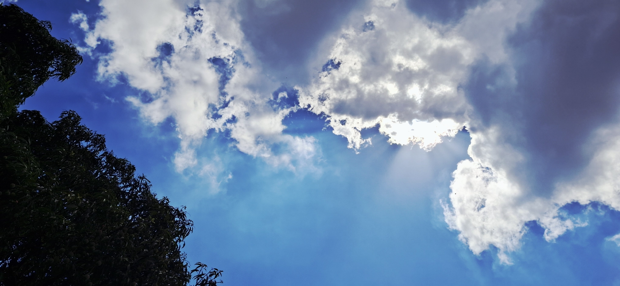 The image shows a view looking up at the sky, with trees visible on the left side of the frame. The sky is primarily blue, with patches of white and gray clouds scattered throughout. Sunlight is filtering through the clouds, creating visible rays of light. The location is Vallecito, Venezuela.