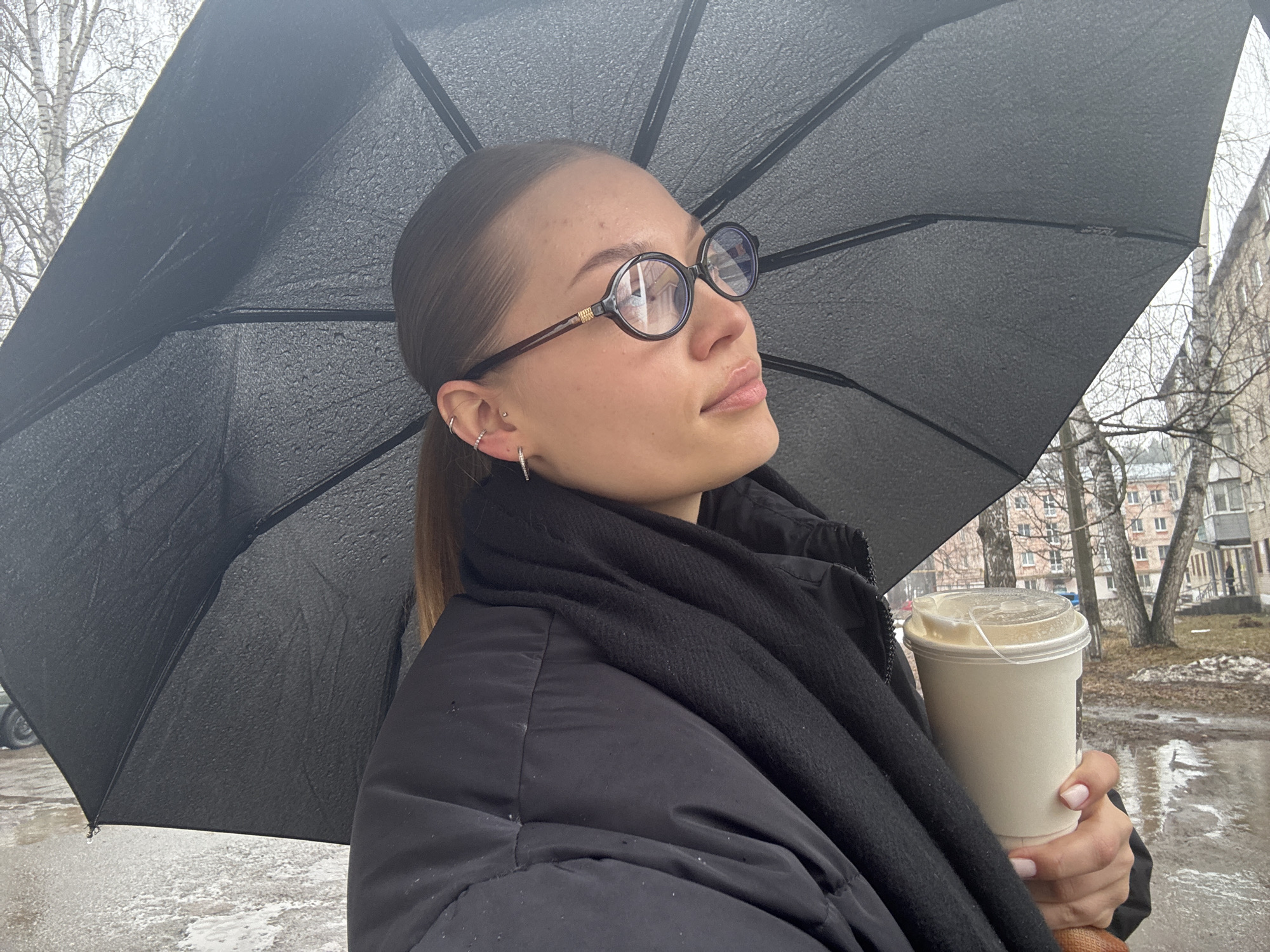 The photograph shows a young woman standing under a black umbrella on a day with some remaining snow, likely in the city of Chaikovsky, Russia. The woman is fair-skinned with brown hair pulled back into a ponytail. She is wearing round eyeglasses and has multiple piercings in her left ear. She has on a black puffy jacket and a black scarf. She is holding a white disposable coffee cup with a clear lid. The background shows a street with bare trees and a multistory residential building. The ground appears wet, indicating recent or ongoing rain or snowmelt.