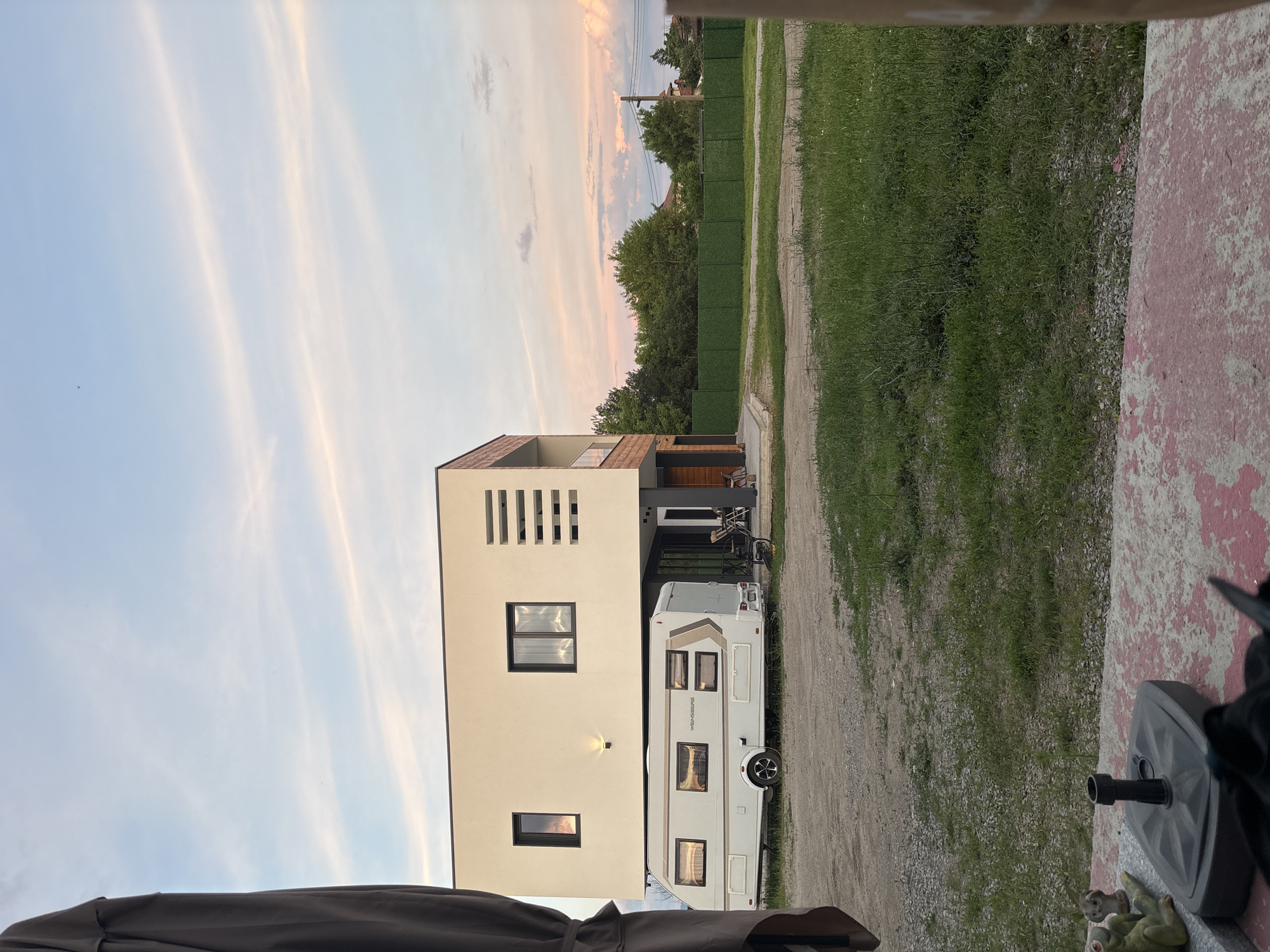 The image depicts a modern, multi-story house with a caravan parked underneath its elevated portion. The house has a light-colored facade and dark-framed windows, with some horizontal slats on the upper story. The caravan is white with brown and grey accents. In front of the house, there is a gravel area, and further in the foreground, a patch of green grass. Beyond the house, a line of trees forms a backdrop, with a green fence running along the property line. The sky is partly cloudy with streaks of pink and orange, suggesting it is either sunrise or sunset. A dark curtain or fabric is visible in the far left of the frame, obscuring part of the view. The scene appears to be in a semi-rural setting, potentially on the outskirts of Mramor, Bulgaria, given the description provided. There are no people or animals clearly visible in the image, and no specific actions or interactions are taking place. The overall impression is one of a residential property with a caravan.