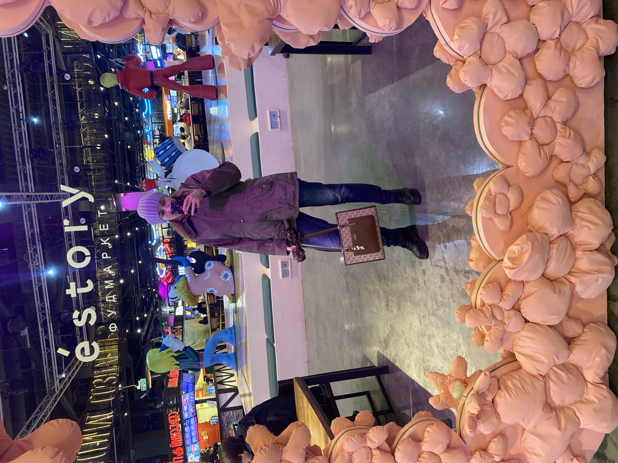 A woman stands facing the camera in front of a photo booth or decorative structure. She is wearing a lavender hat, a dark-colored jacket, blue jeans, and black boots. She is holding a purse in her right hand and has a face mask pulled down to her chin.
The structure surrounding her appears to be a frame adorned with pink, puffy flower-like shapes.
The background shows the interior of a food market or food court. There are various food stalls and decorative elements visible, including large cartoon character figures. The market has a modern industrial feel with exposed ceilings and lighting fixtures.
Based on the signage visible in the background, the location is "EStory Food Market" in Yekaterinburg, Russia. There are also tables and chairs nearby, suggesting a seating area for the food market. Other people are present in the background near the food stalls.