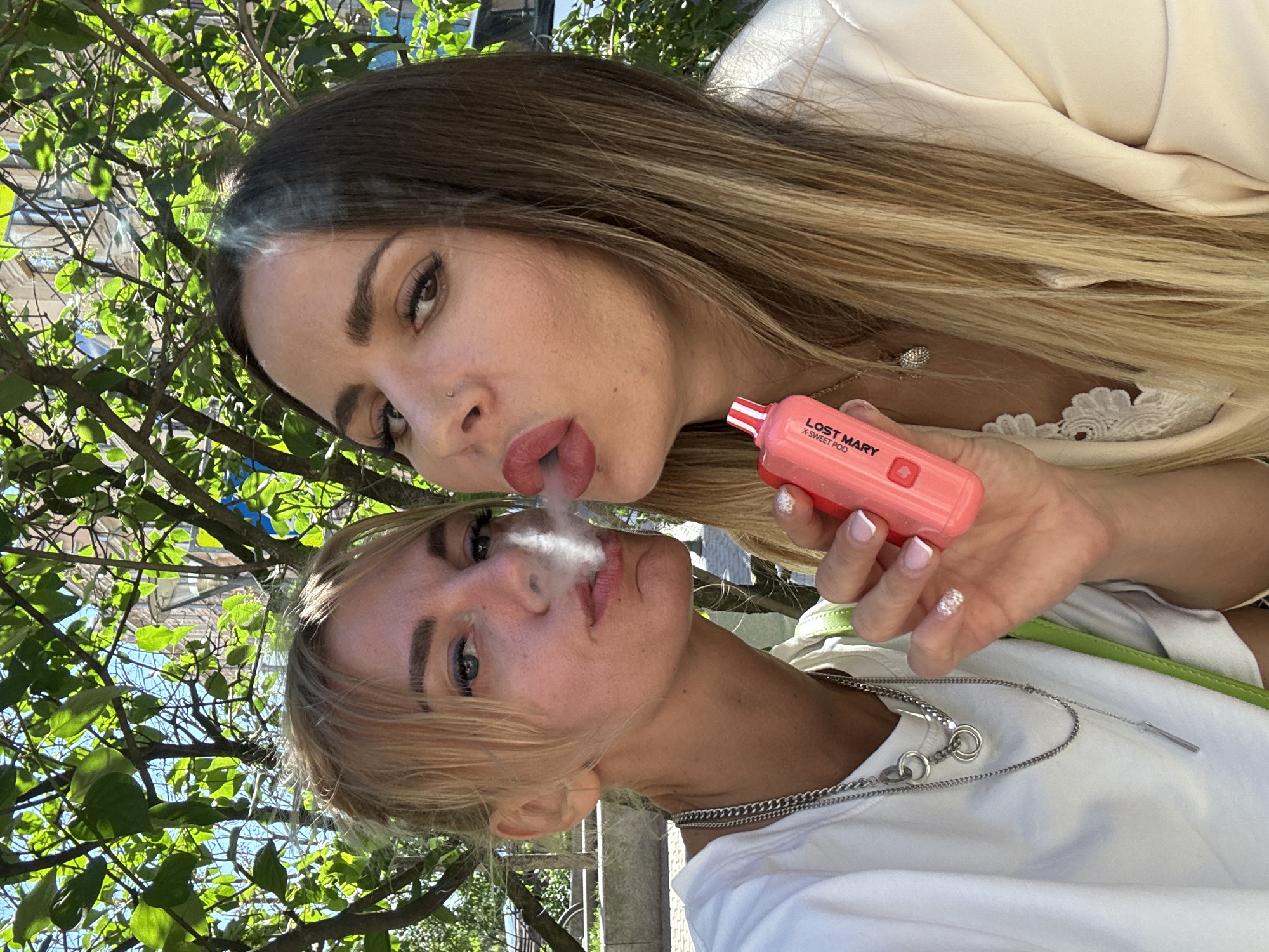 This is a vertical, close-up selfie of two young women outdoors. The woman in the foreground has blonde hair and is looking towards the camera, with smoke exhaling from her mouth. She is wearing a white t-shirt and a silver chain necklace. The woman behind her, with long brown hair, is also exhaling smoke. She is holding a pink electronic cigarette, which has "LOST MARY X-SWEET POD" printed on it. She is wearing a light-colored top with lace detailing. The background shows green foliage and blue sky, suggesting a daytime setting. The image appears to be taken on a sunny day. The women's expressions are difficult to discern due to the close-up nature of the shot and the smoke, but they seem relaxed. The overall vibe is casual and candid. The mention of Saint Petersburg, Russia, places the scene in an urban environment with likely pleasant weather for outdoor activities.