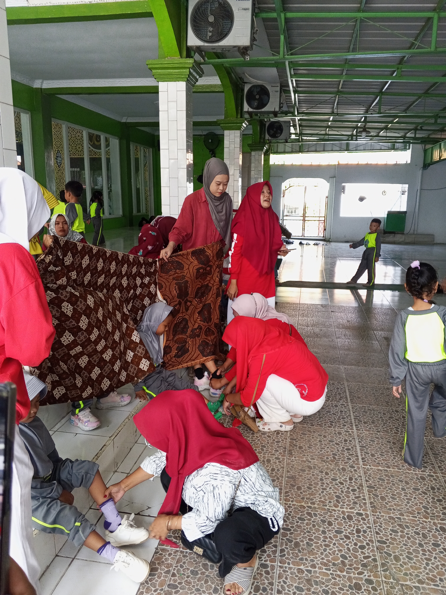 The image depicts an indoor scene, likely a community hall or mosque, in Gebang, Indonesia, characterized by green and white pillars, a green ceiling with exposed beams and metal roofing, and air conditioning units mounted high on the walls. The floor is tiled with a mosaic of brown and tan stones. Several people, predominantly children and some adults wearing hijabs, are engaged in activities.
In the foreground, a child in a red hijab and white pants is being attended to by an adult wearing a similar red hijab and a patterned white and black shirt. The adult is helping the child put on white sneakers over purple socks. To the left of this interaction, another child in a gray outfit and red hijab is partially visible.
Behind them, a large patterned brown and white fabric is spread out. Several children are interacting with this fabric, some sitting on it, and one child in a gray and yellow uniform is partially hidden underneath it. An adult in a red hijab stands to the right, holding one edge of the fabric.
Further in the background, a child in a green and gray uniform is standing and facing away from the camera, with one arm extended. Another child in a similar gray and yellow outfit is walking away towards the right. The environment appears to be a gathering, possibly for an event or lesson, with children and adults interacting in a relaxed manner within the covered space.