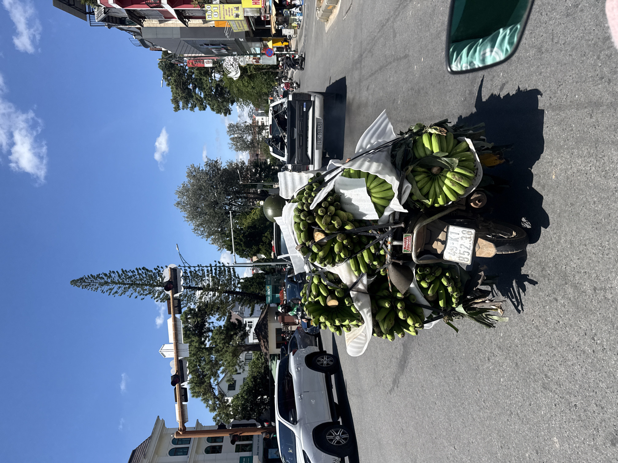 A daylight, eye-level shot captures a motorcycle laden with numerous bunches of green bananas, traveling on an asphalt road in Da Lat, Vietnam. The motorcycle is in the foreground, with the bananas wrapped in white fabric and secured with black straps, overflowing from the back. A license plate with the text "49-K1 852.38" is visible on the rear of the motorcycle. Behind the motorcycle, a white Ford F-150 pickup truck is in motion, its license plate reading "49C-169.45". In the background, buildings with European architectural styles, trees, and traffic lights line the street under a bright blue sky with scattered clouds. A white SUV is parked on the left side of the road, and other motorcycles and pedestrians are visible further down the street. The scene appears to be a busy city intersection.