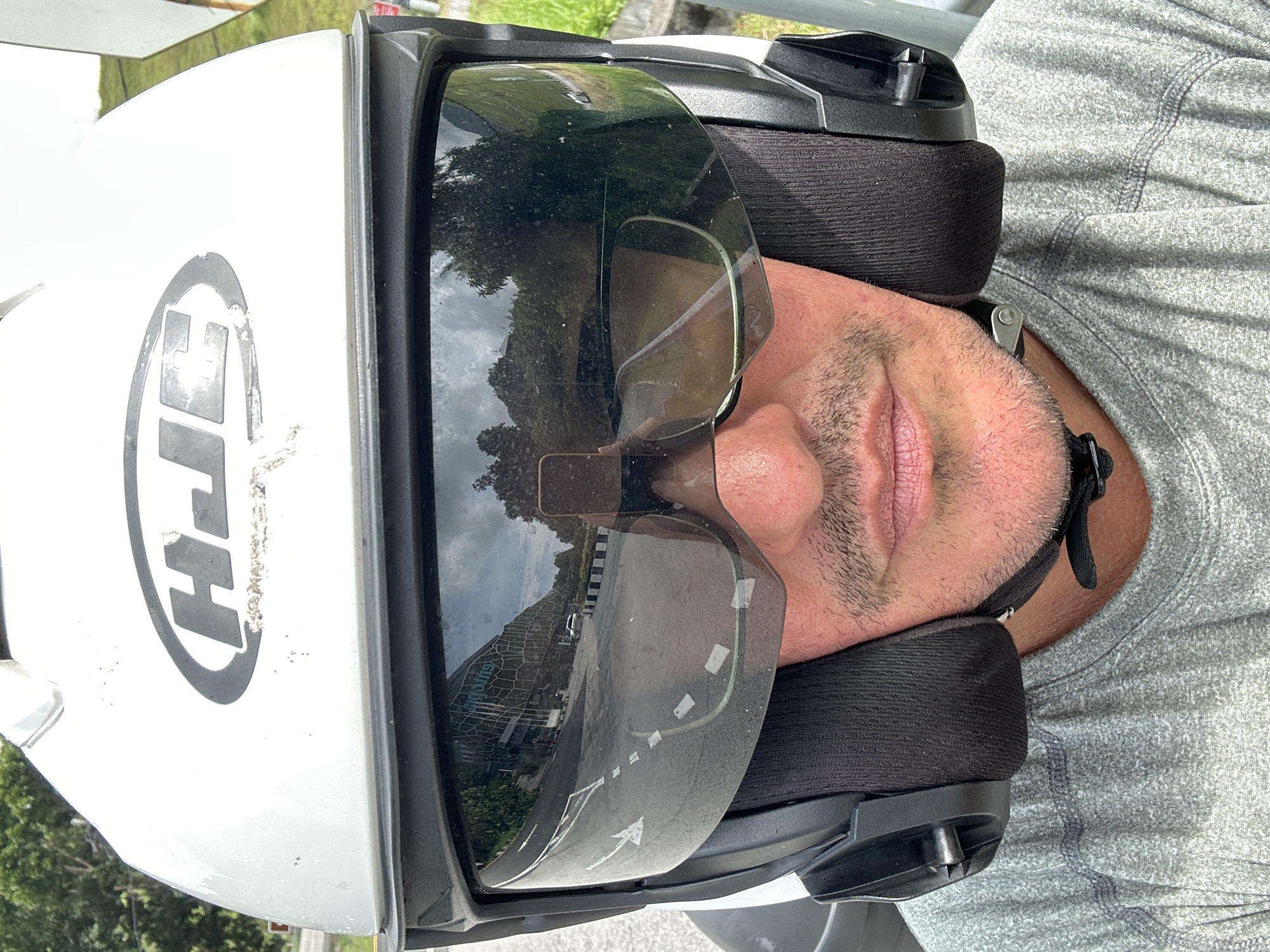 The image shows a close-up, selfie-style view of a person wearing a white HJC motorcycle helmet and a grey t-shirt. The helmet has a dark tinted visor that reflects a road, trees, a cloudy sky, a building, and a white car. The person's face, including their mouth and chin stubble, is visible. The background suggests an outdoor environment with greenery and what appears to be a road and possibly a building or complex. The reflection in the visor indicates the person is likely in an urban or semi-urban setting. Given the context of George Town, Malaysia, this could be a street scene within or near the city.