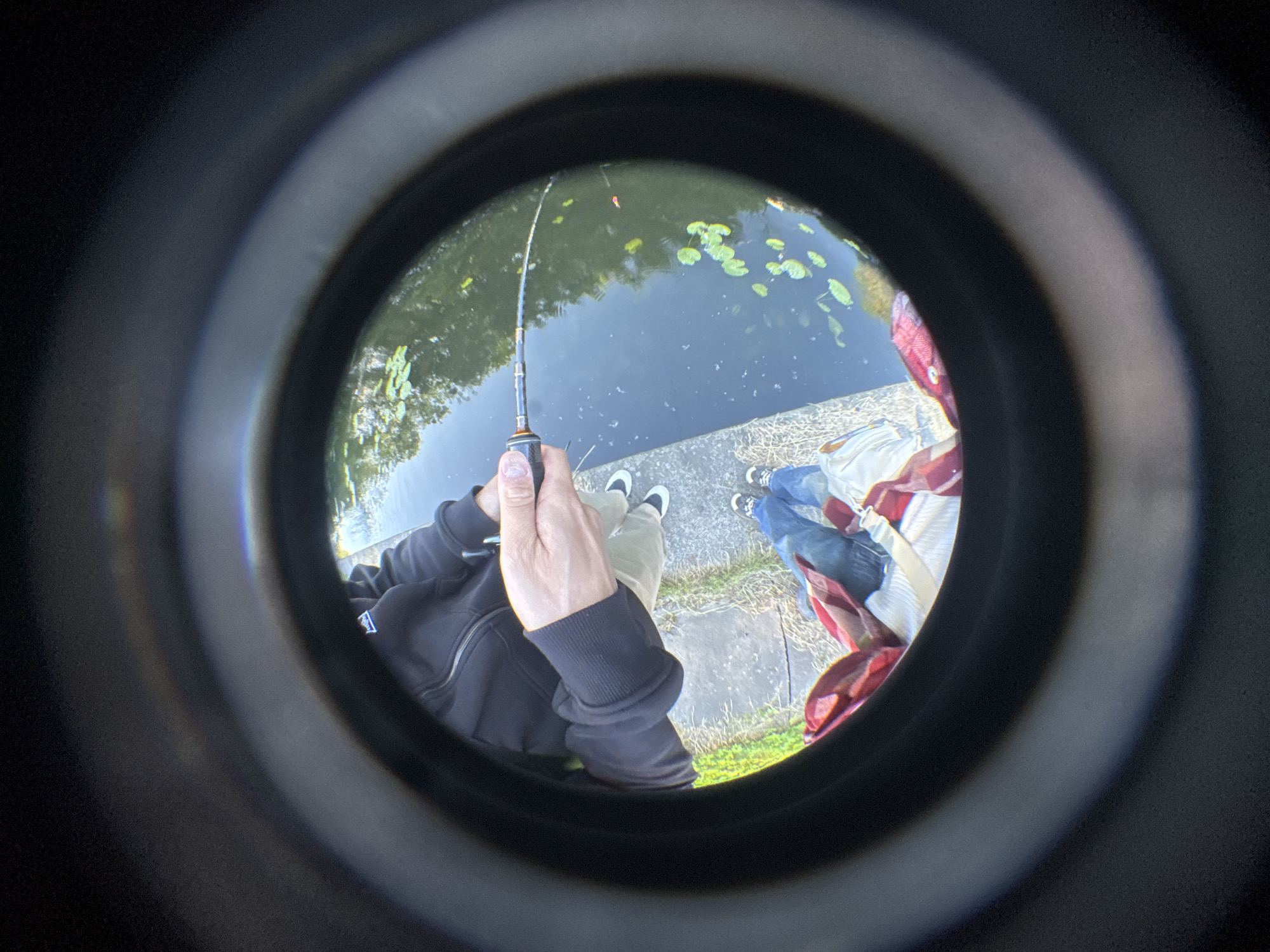 The image, taken through a circular lens, shows a person fishing by a body of water. The person is wearing a black hooded jacket, tan pants, and sneakers. They are holding a fishing rod over the water, which has lily pads and reflections of trees. The scene appears to be a canal or riverbank. The overall effect is somewhat distorted due to the lens.