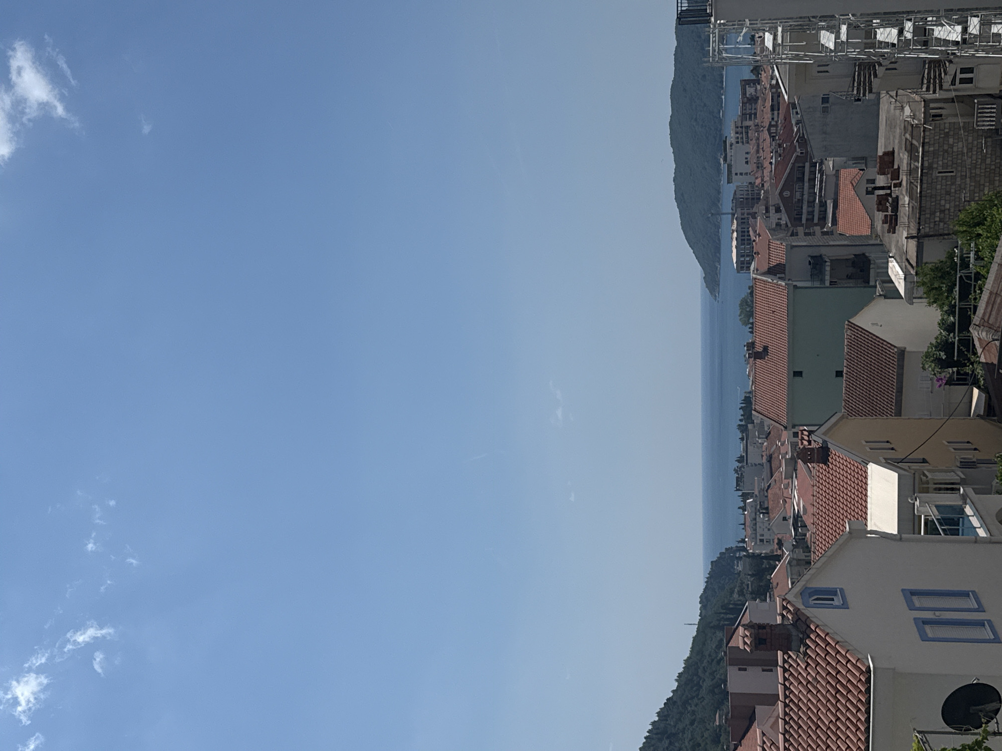 This is a distant, high-angle, medium shot of a coastal town in Budva, Montenegro, on a clear day with a few wispy clouds. The foreground is dominated by the terracotta tiled roofs of residential buildings. Some of these buildings have white or light-colored facades, and a few feature distinctive blue window frames.
The middle ground showcases a densely packed urban landscape with more buildings of varying heights and architectural styles, also with predominantly red-tiled roofs. Beyond the town, a large expanse of the Adriatic Sea is visible, with a distant, green, mountainous island or peninsula on the right side of the frame. Another, smaller green hillside is present on the left, bordering the town. The sky is a bright, clear blue. No people are visible in the frame. The overall impression is one of a sun-drenched Mediterranean settlement.