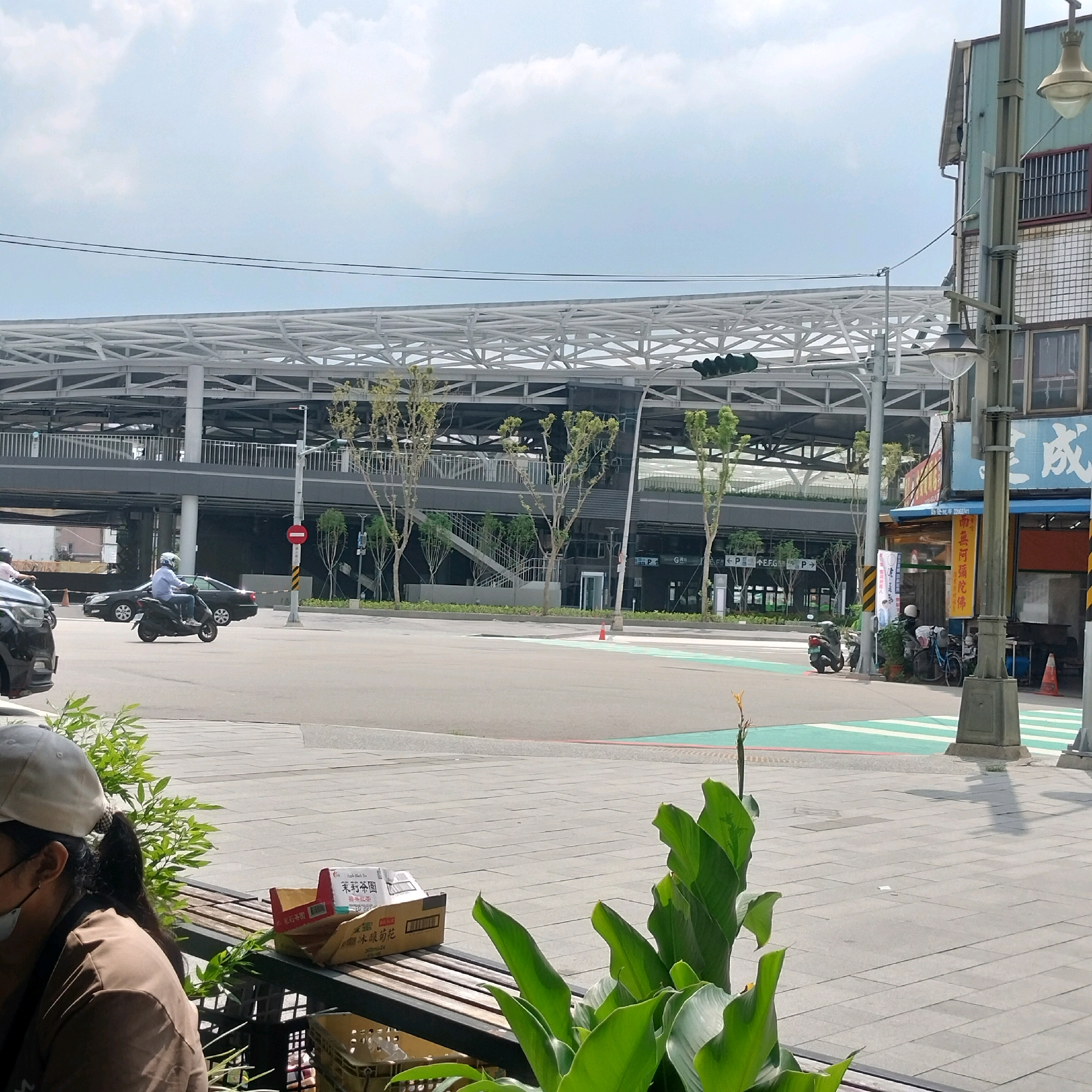 The image captures a street scene in Taichung, Taiwan, likely near a transportation hub. In the foreground, a person wearing a light-colored baseball cap and a brown top is seated on a bench, partially obscured by lush green foliage. Next to them on the bench are two cardboard boxes, one of which is open.
The middle ground features a street intersection with a pedestrian crossing marked by green and white stripes. A motorcycle rider wearing a white helmet and a light blue shirt is on a scooter, navigating the intersection. Behind the scooter, a black sedan is visible. In the background, a modern, multi-level elevated structure, possibly a train station or elevated highway, dominates the skyline. The structure is characterized by a white, lattice-like roof and dark supporting elements.
Further in the background, to the right, is a building with a green metal facade and windows with grates. Signs with Chinese characters are attached to the building. Several trees with sparse foliage are planted along the walkway and around the elevated structure. Traffic lights are visible on poles, and a red circular sign with a white bar is mounted on a pole near the intersection. The sky is partly cloudy.