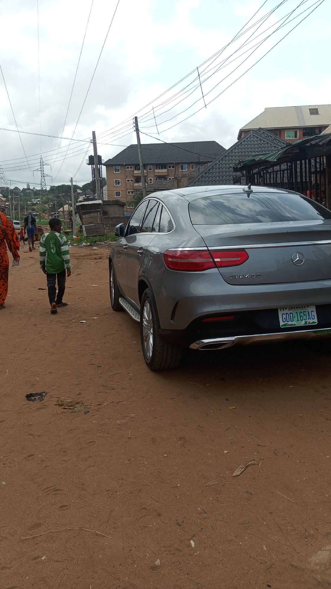 The image captures a scene on a dirt road in Nkpor, Nigeria. A gray Mercedes-Benz GLE 400 is parked on the right side of the road, its rear facing the camera. The license plate reads "GDD-165AG" and indicates the state of Anambra. A young boy wearing a green and white striped shirt and black pants walks along the left side of the road, moving away from the camera. A person in an orange patterned outfit is partially visible on the left edge of the frame. In the background, buildings and electrical poles are visible against a cloudy sky. Debris is scattered on the dirt road.