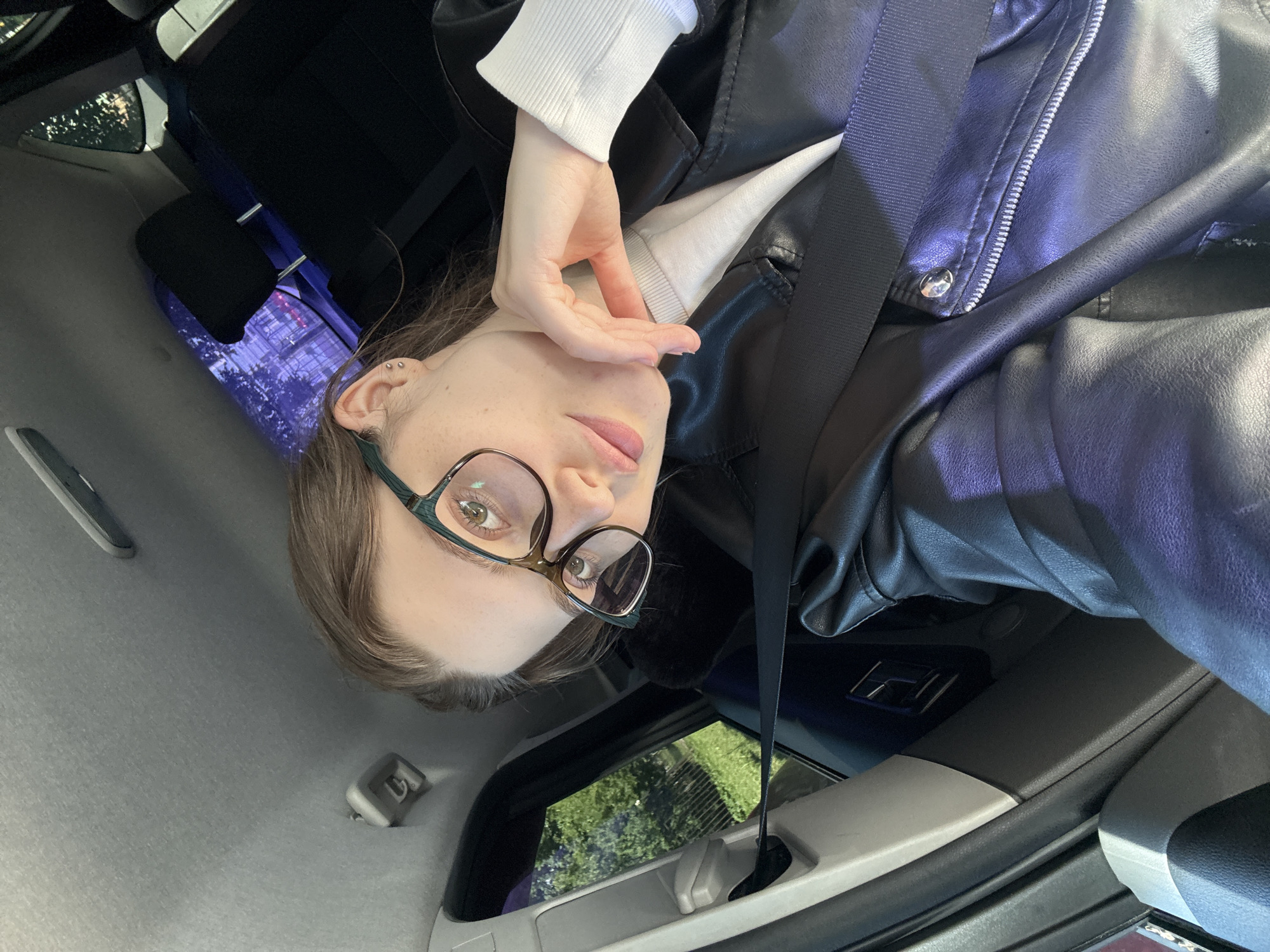 A young woman with light brown hair and glasses is in the back seat of a car. She is wearing a white sweater and a black leather jacket. Her left hand is resting on her chin. The interior of the car is visible, including a seatbelt and the car's ceiling. Outside the window, greenery and trees can be seen, suggesting a suburban or park-like setting within or on the outskirts of Saint Petersburg, Russia. The lighting indicates it is daytime.