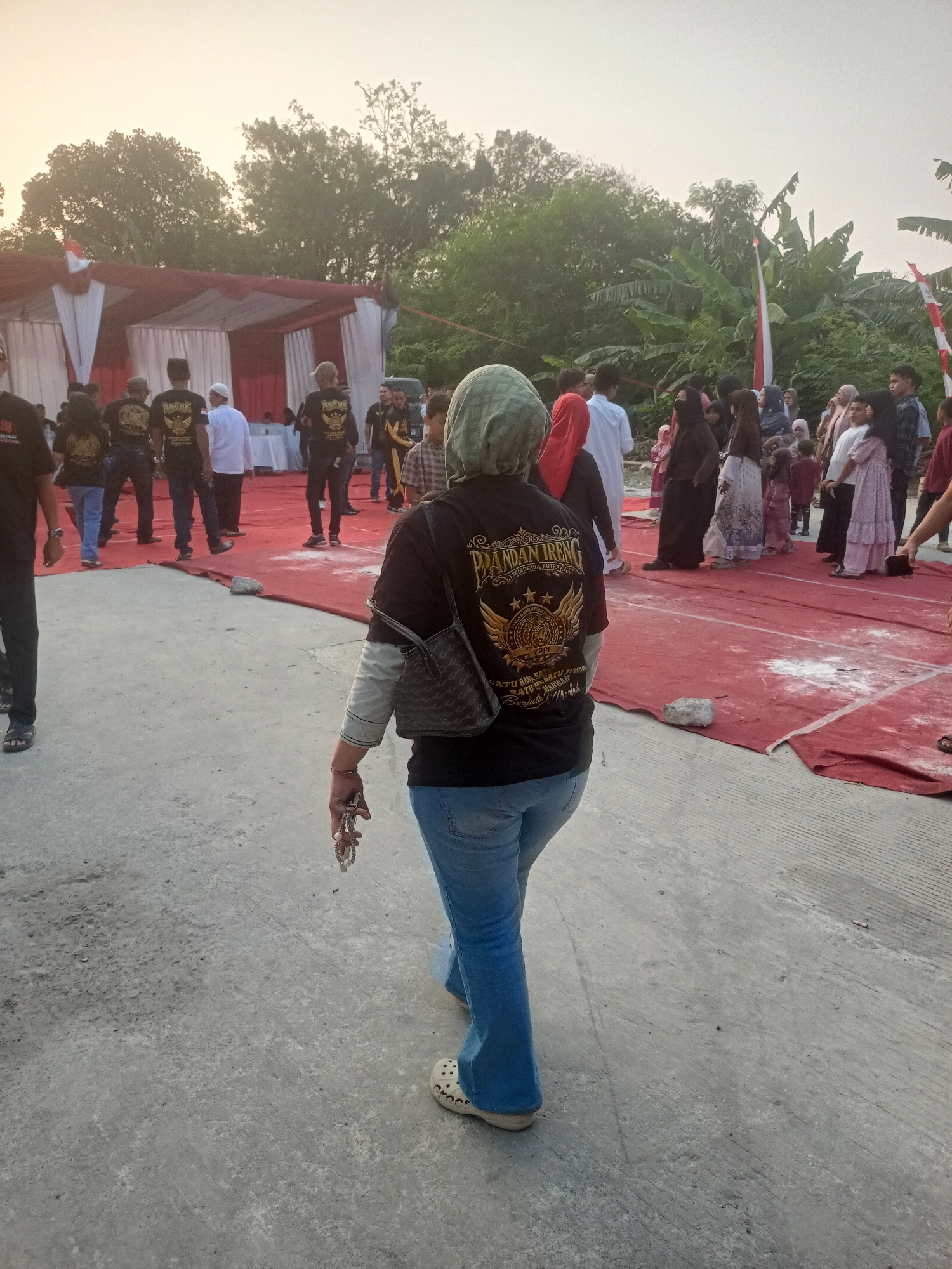 In Bekasi, Indonesia, a gathering is taking place outdoors, likely for an event given the red carpet laid out and the tents in the background. A woman wearing a light green hijab and blue jeans walks away from the camera, her back towards us. She carries a dark handbag over her left shoulder and holds a bracelet in her right hand. She is barefoot with white clogs on. Behind her, a crowd of people are standing, many wearing black t-shirts with a gold emblem and text. Some of these t-shirts have "PANDAN IRENG" and other text visible. The ground is a mix of concrete and red carpet, with some white powder scattered on the carpet. The environment includes trees and lush vegetation, suggesting a location on the outskirts of the city or a park. The lighting indicates it is either early morning or late afternoon.
