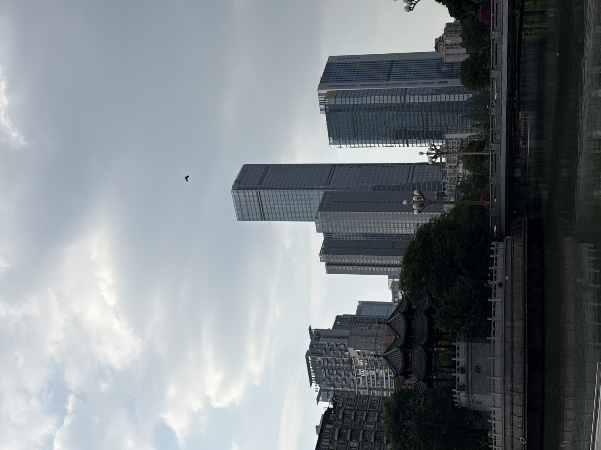 An outdoor, eye-level shot captures a cityscape in Wuhou District, China, under an overcast sky with visible clouds. The upper half of the image is dominated by a light grey sky, where a small, dark bird is visible in flight in the upper-center left quadrant. The background features a cluster of modern, high-rise buildings, predominantly dark-toned and constructed with reflective glass and concrete.
In the midground, a traditional Chinese pavilion with a dark, multi-tiered roof and intricate architectural details stands on a light grey stone platform on the left bank of a body of water, likely a river or canal. Dense, dark green trees and foliage surround this pavilion and extend along the banks. A grey stone bridge with multiple ornate, multi-bulb lampposts crosses the water, connecting the foreground to the midground on the right. The water in the foreground and midground is dark and calm, reflecting the overhead sky and some elements of the architecture and vegetation. No identifiable people are present.
