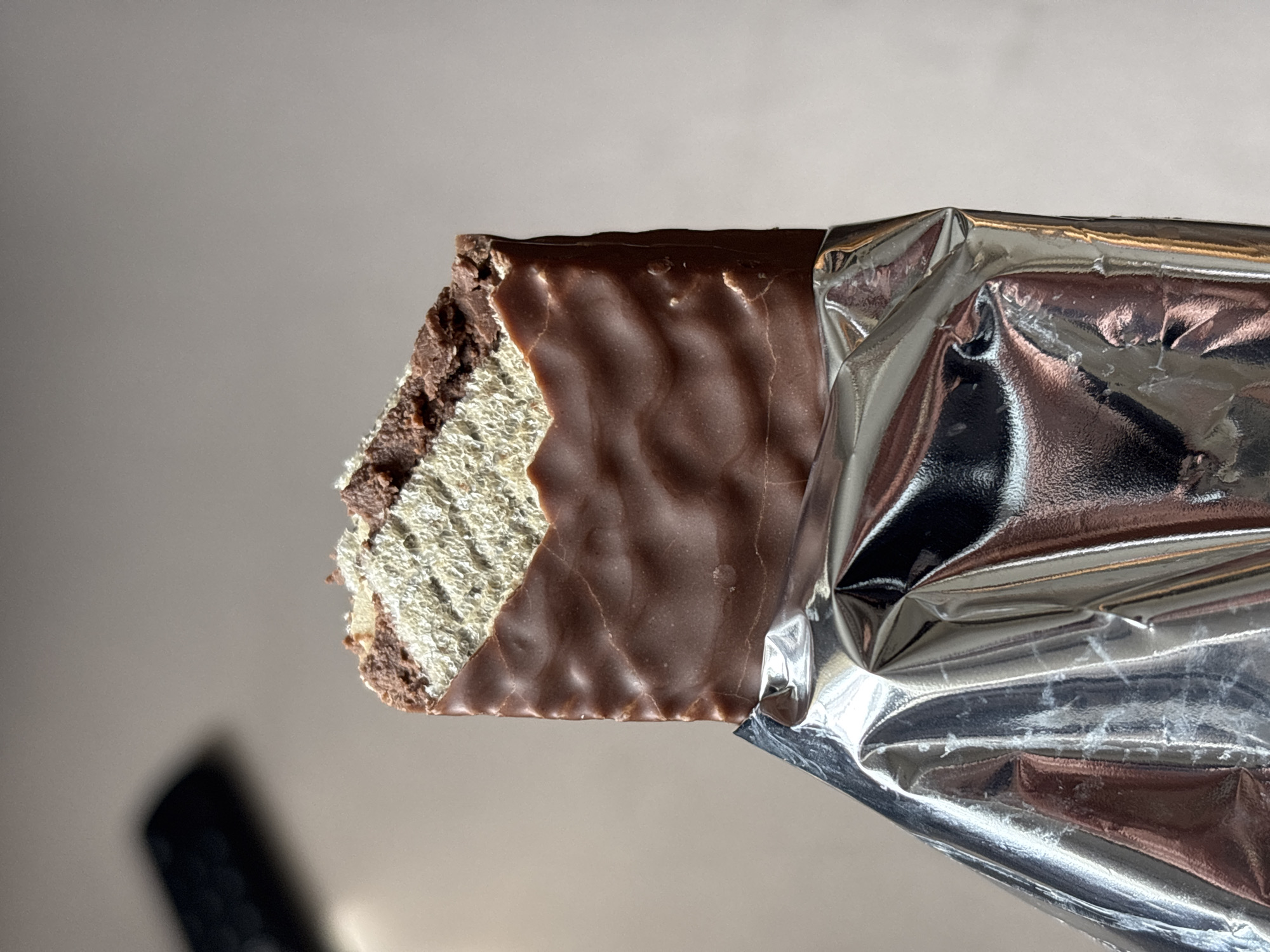 A partially unwrapped chocolate-covered wafer bar is prominently displayed. The chocolate coating is dark brown and uneven, indicating it has been bitten into on the left side. Inside, the wafer layers are visible, appearing light in color. The wrapper is silver and crumpled, with a portion peeled back to reveal the chocolate bar. The background is a plain, light color. A dark, blurry shape is visible in the lower left corner, which could be another object or shadow.
