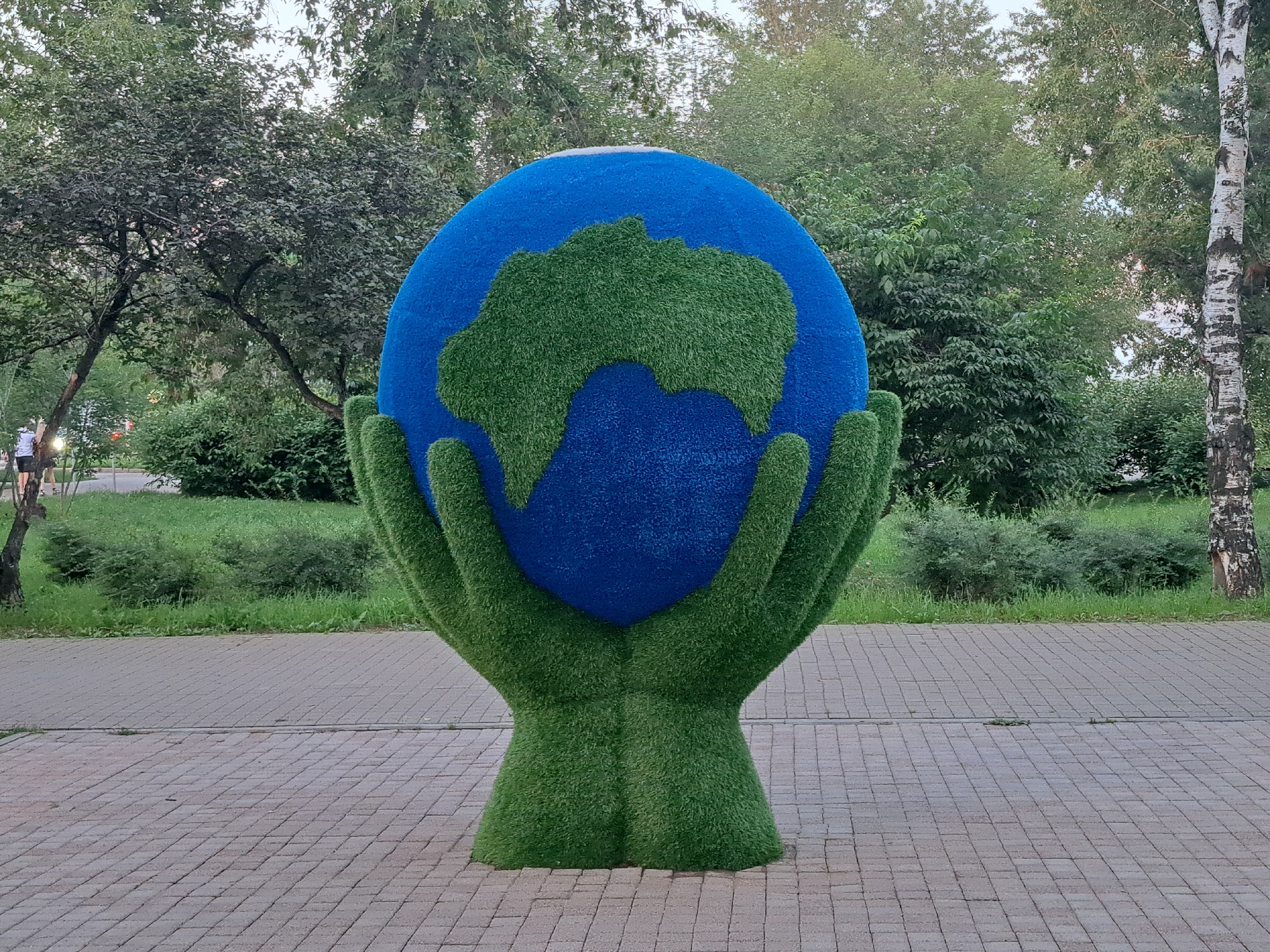 The image depicts a sculpture of the Earth held by two grass-covered hands, situated on a paved pathway in a park-like setting. The Earth is represented by a blue sphere with green continents. The sculpture is positioned outdoors, with lush green trees and bushes forming the background. A birch tree with distinctive black and white bark is visible to the right. The pathway is paved with light-colored rectangular stones. In the distance, a few indistinct figures can be seen walking. The lighting suggests it is daytime, possibly late afternoon or early evening, with a slightly overcast sky. The scene is static, with no apparent activity or interaction occurring. The sculpture appears to be an art installation or a decorative element in a public space.