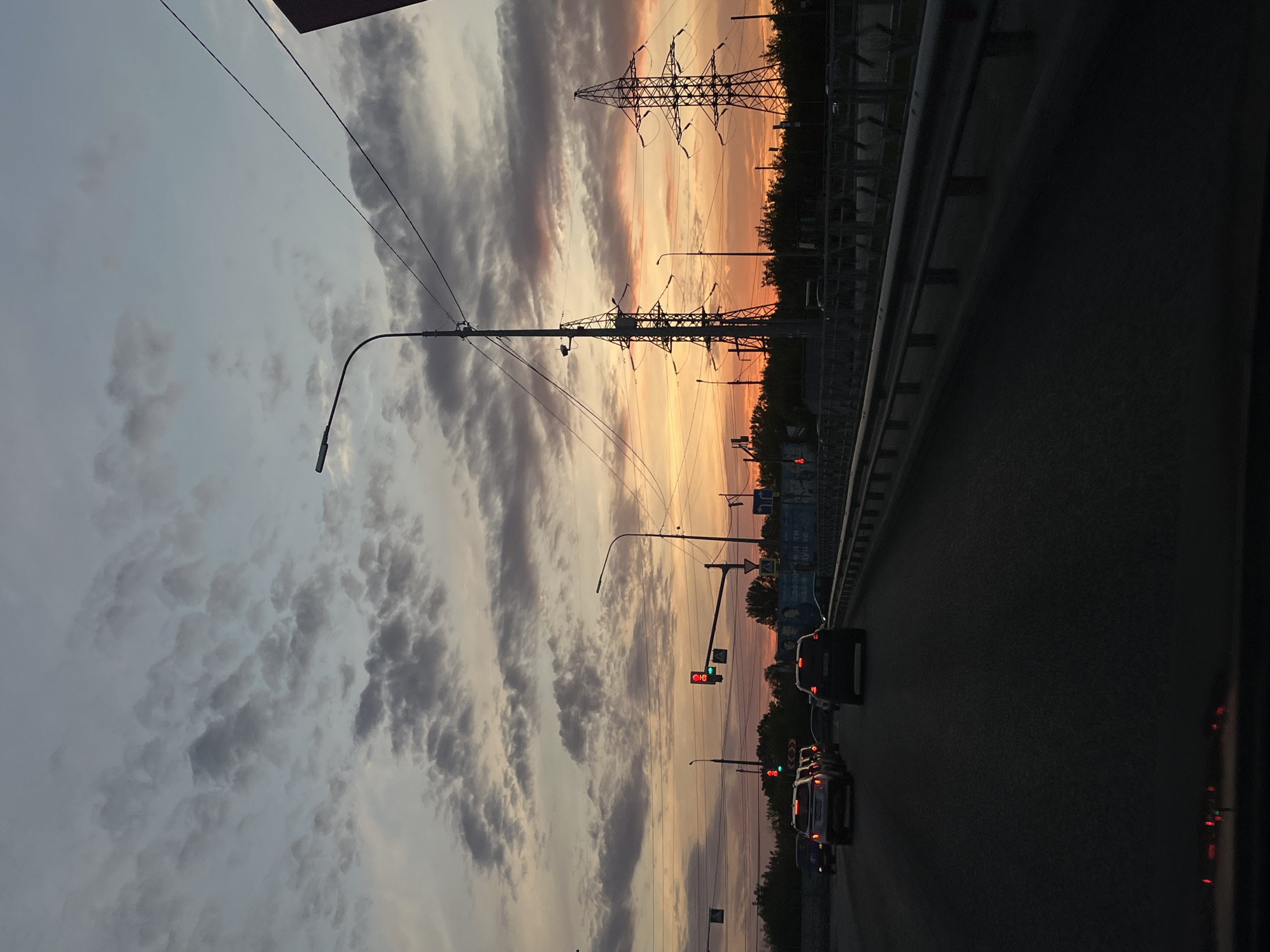 The image captures a sunset over a highway in Novokuybyshevsk, Russia. The sky is a canvas of soft grays and oranges, with scattered clouds. Power lines and towering electricity pylons dominate the silhouette against the fading light. Several cars are visible on the road, indicating a moderate flow of traffic. Traffic lights are present, with one showing a red signal. Trees line the side of the highway, and a metal guardrail runs along the road's edge. The foreground is dominated by the dark asphalt of the road, with faint red lights reflecting from within what appears to be a vehicle.