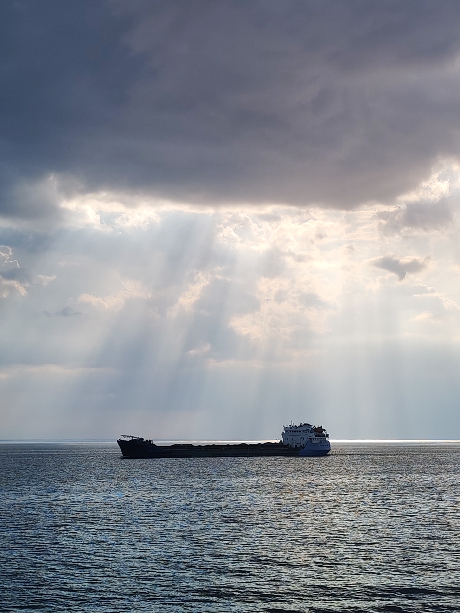 A cargo ship is on the water under a cloudy sky with light rays shining through in an unknown location in Russia. The ship is long and low in the water, with a dark hull and a lighter-colored superstructure at one end. The water is choppy, with a slight sheen. The sky is dominated by heavy, dark clouds, but bright light is breaking through in several places, creating beams of light that illuminate the water below.
