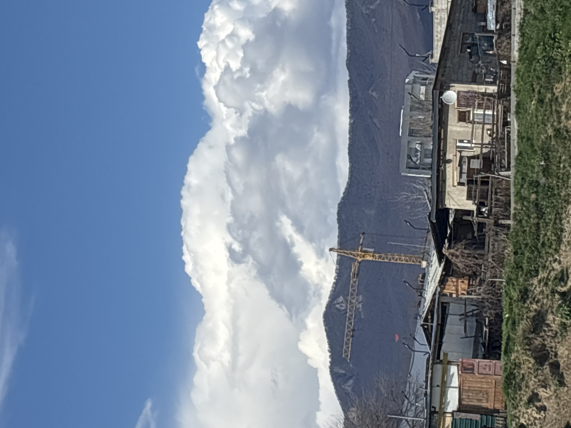 The image depicts a scene in Tbilisi, Georgia, with a mountain backdrop. Large, fluffy white clouds dominate the left side of the frame, contrasting with a clear blue sky. A mountain range runs vertically in the middle of the image. A yellow construction crane is visible against the mountain. To the right, there's a cluster of buildings, appearing somewhat dilapidated. Green vegetation is visible in the extreme right foreground. A satellite dish is mounted on one of the buildings.