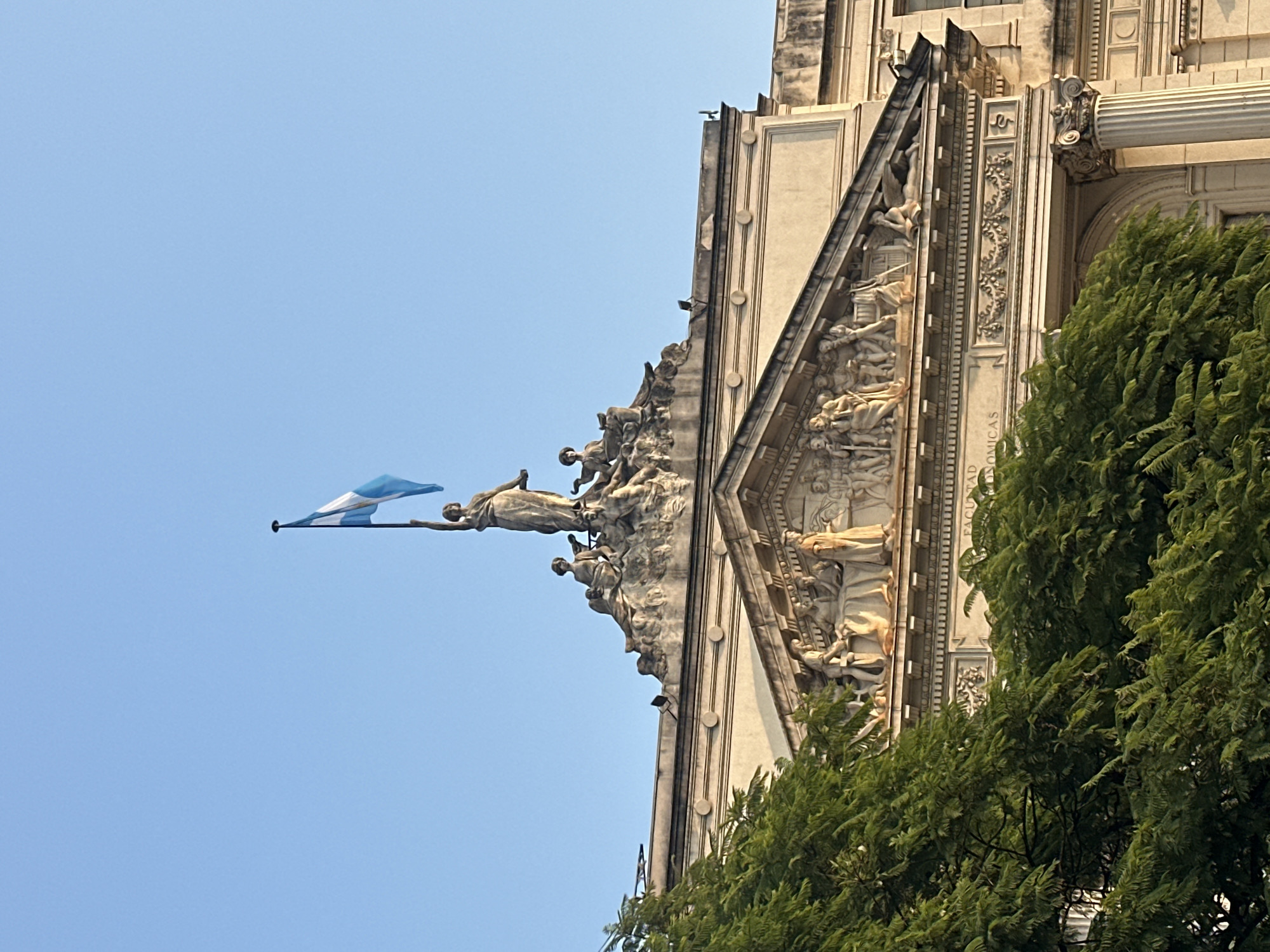 A low-angle shot shows a portion of an ornate building facade in Buenos Aires, Argentina, likely the Law School of the University of Buenos Aires. The building is of a classical style with multiple figures sculpted along its upper edges and along a triangular pediment. The Argentine flag is seen waving on the upper left of the building, with a figure appearing to hold the flag pole. The sky is clear blue. A portion of a leafy green tree is seen on the right side of the image, partially obscuring the lower portion of the building. Architectural detailing includes a column with an ornate capital and carved designs with scrolling foliage. The light is bright and suggests daytime.