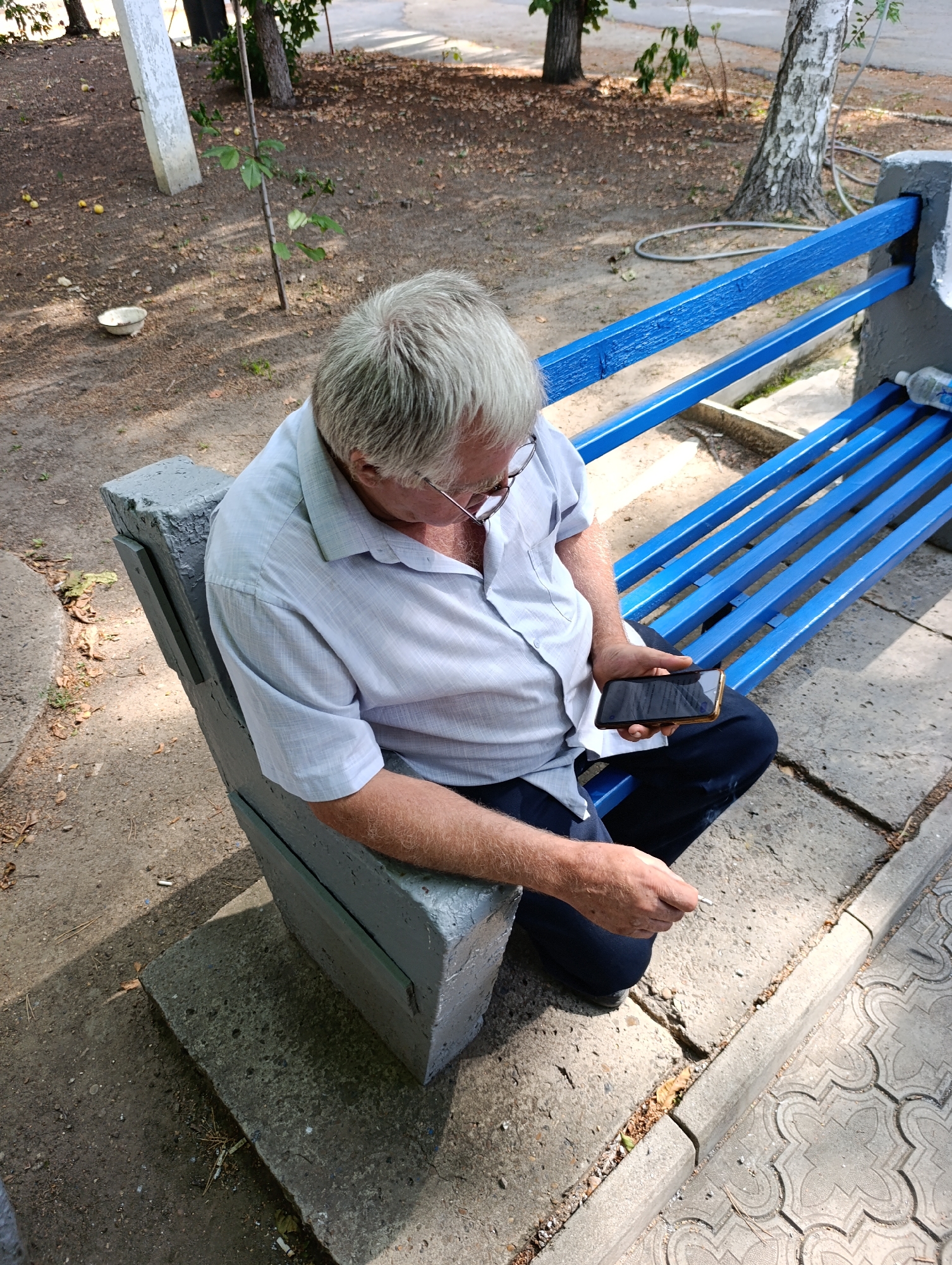 An elderly man with graying hair and glasses sits on a concrete bench with blue wooden slats, holding a smartphone in his right hand. He is wearing a light-colored collared shirt and dark pants. The bench is situated outdoors in an area with trees, fallen leaves, and a paved walkway. The overall setting appears to be a park or public outdoor space, possibly within the Dubăsari town council area of Moldova, given the prompt's context. The lighting suggests it is daytime.