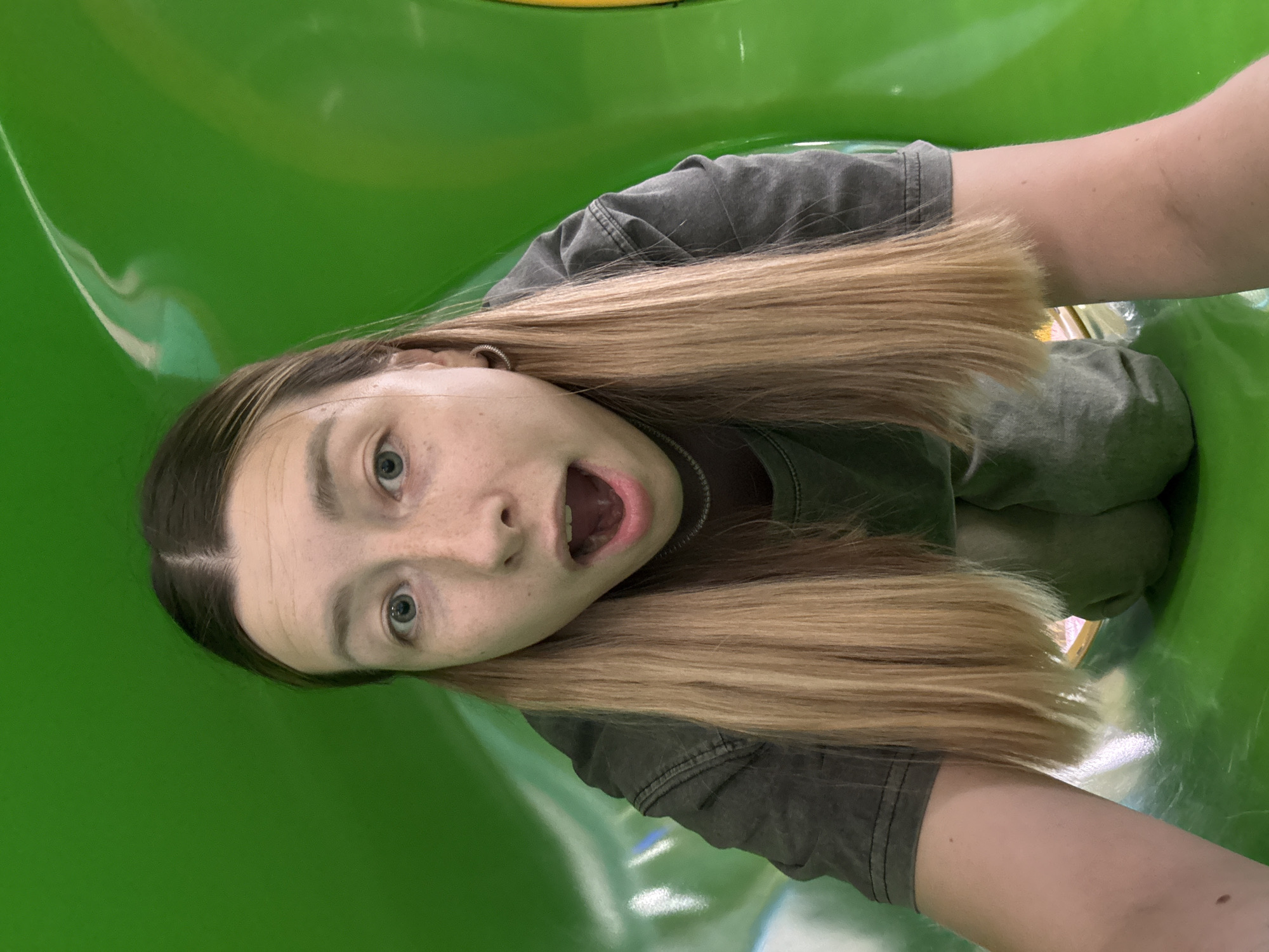 A young woman is pictured inside a green plastic tunnel, possibly a slide. She has fair skin, light brown hair with blonde highlights, and open mouth, looking directly at the camera. She is wearing a dark gray shirt. The interior of the tunnel appears to be brightly lit. The image does not provide information that can be associated with the city of Khimki, Russia.