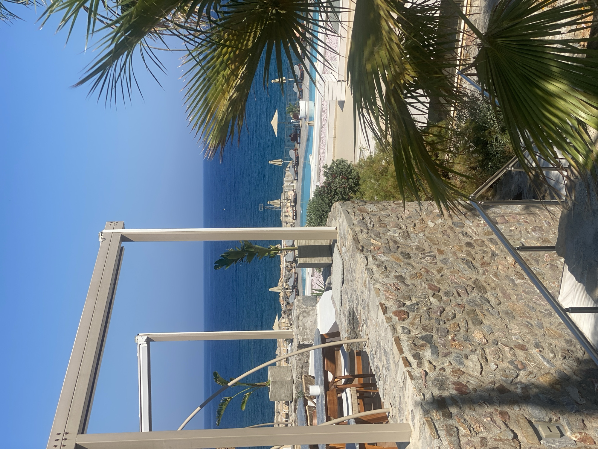 This is a medium-shot, eye-level, exterior photograph taken at a resort in Panormos, Greece. The scene depicts a stone wall in the foreground, leading down a set of stairs with a stainless steel handrail. To the right, palm fronds are visible. Beyond the wall, a swimming pool with lounge chairs and umbrellas is situated, overlooking the bright blue Aegean Sea. The sky is clear and blue. The overall ambiance is tranquil and idyllic.